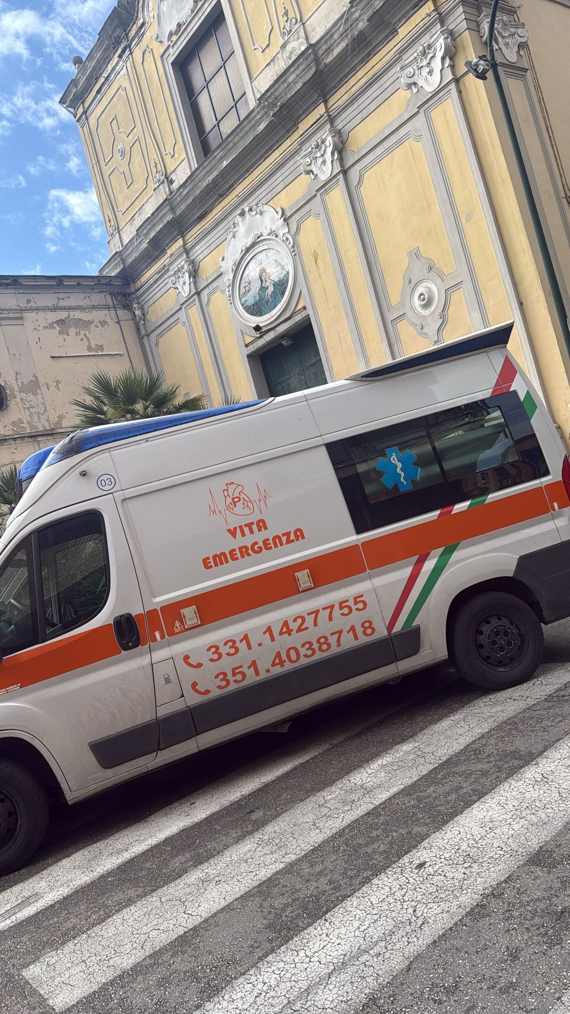 Un furgone di emergenza bianco con segnaletica rossa è parcheggiato sulle strisce pedonali accanto a un edificio storico giallo.