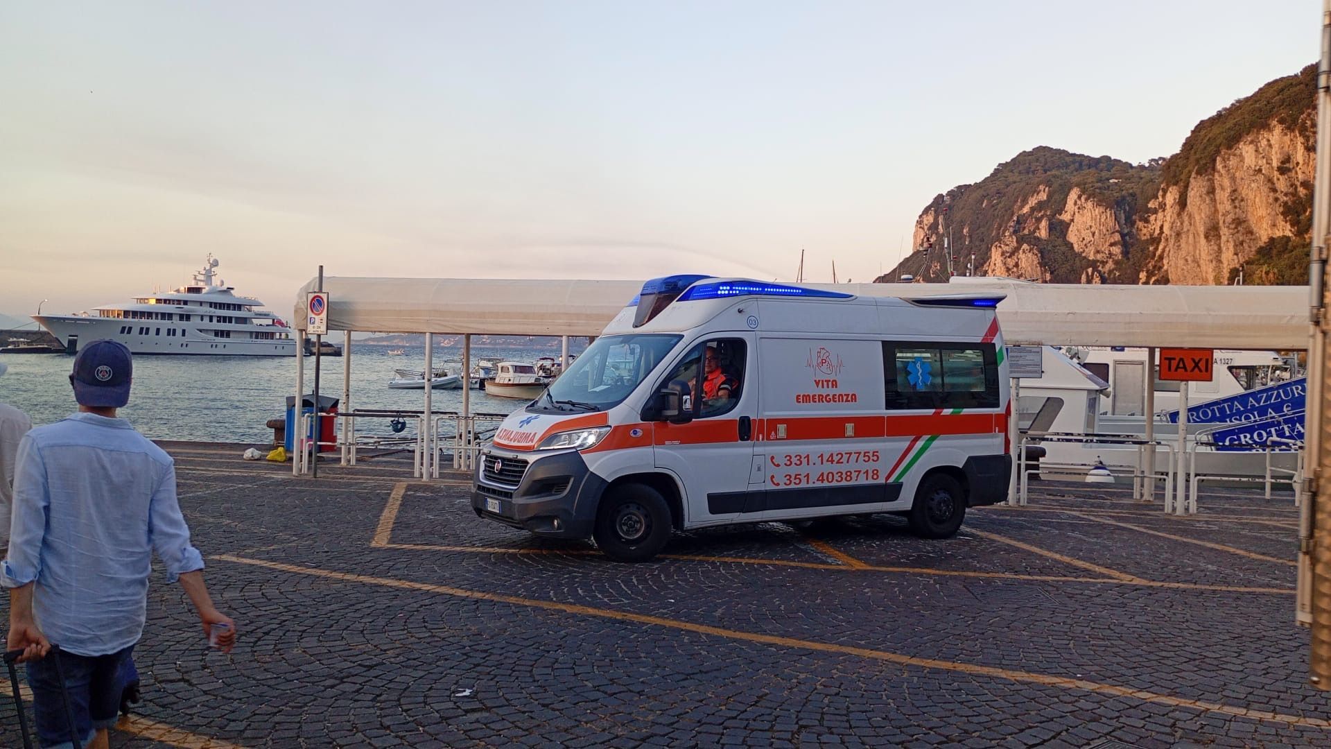 Un'ambulanza parcheggiata sul lungomare al tramonto, con una barca, una scogliera rocciosa e una persona nelle vicinanze.