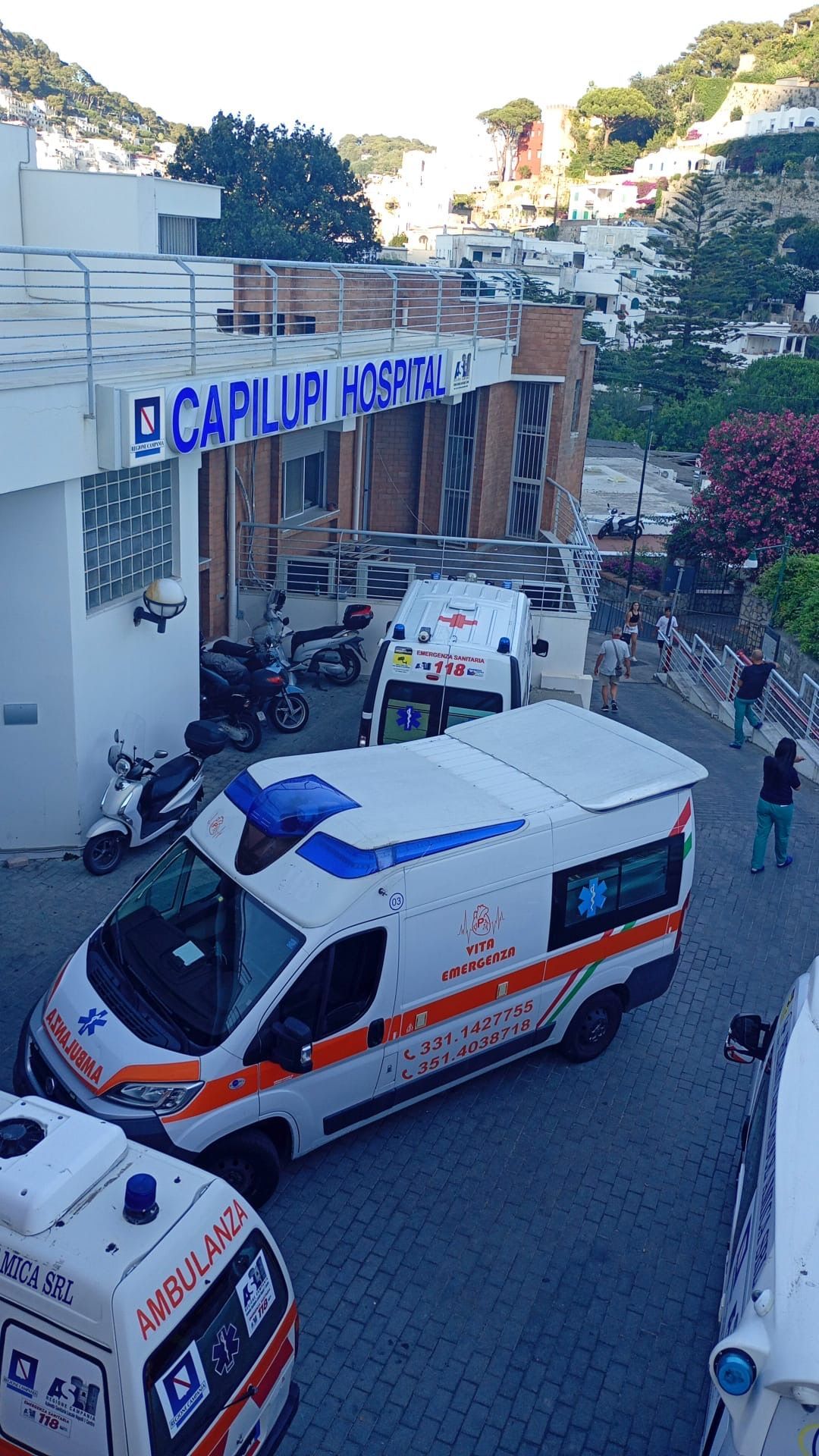 Ambulanza e veicoli di emergenza all'esterno dell'ingresso dell'ospedale di Capilaw, su una strada in salita.