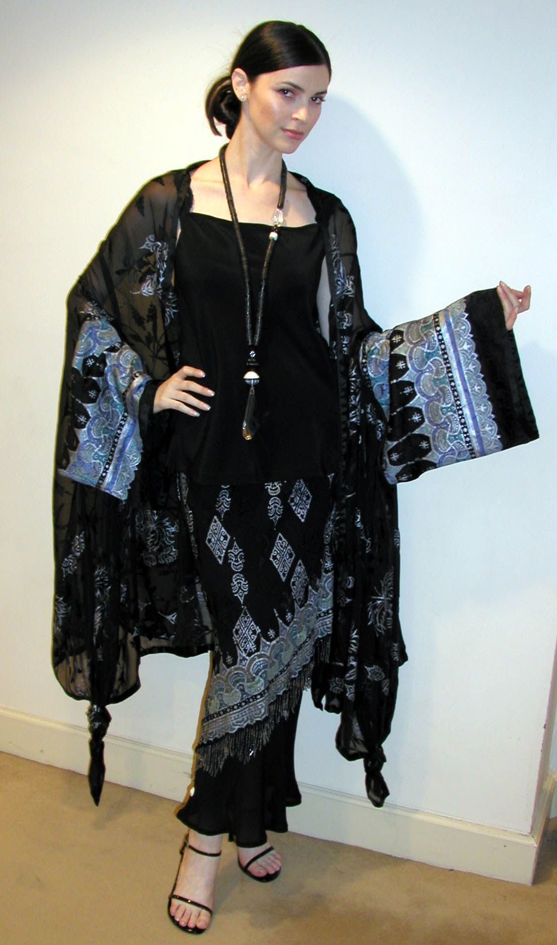 Woman in black outfit with printed shawl and long necklace, posing indoors.