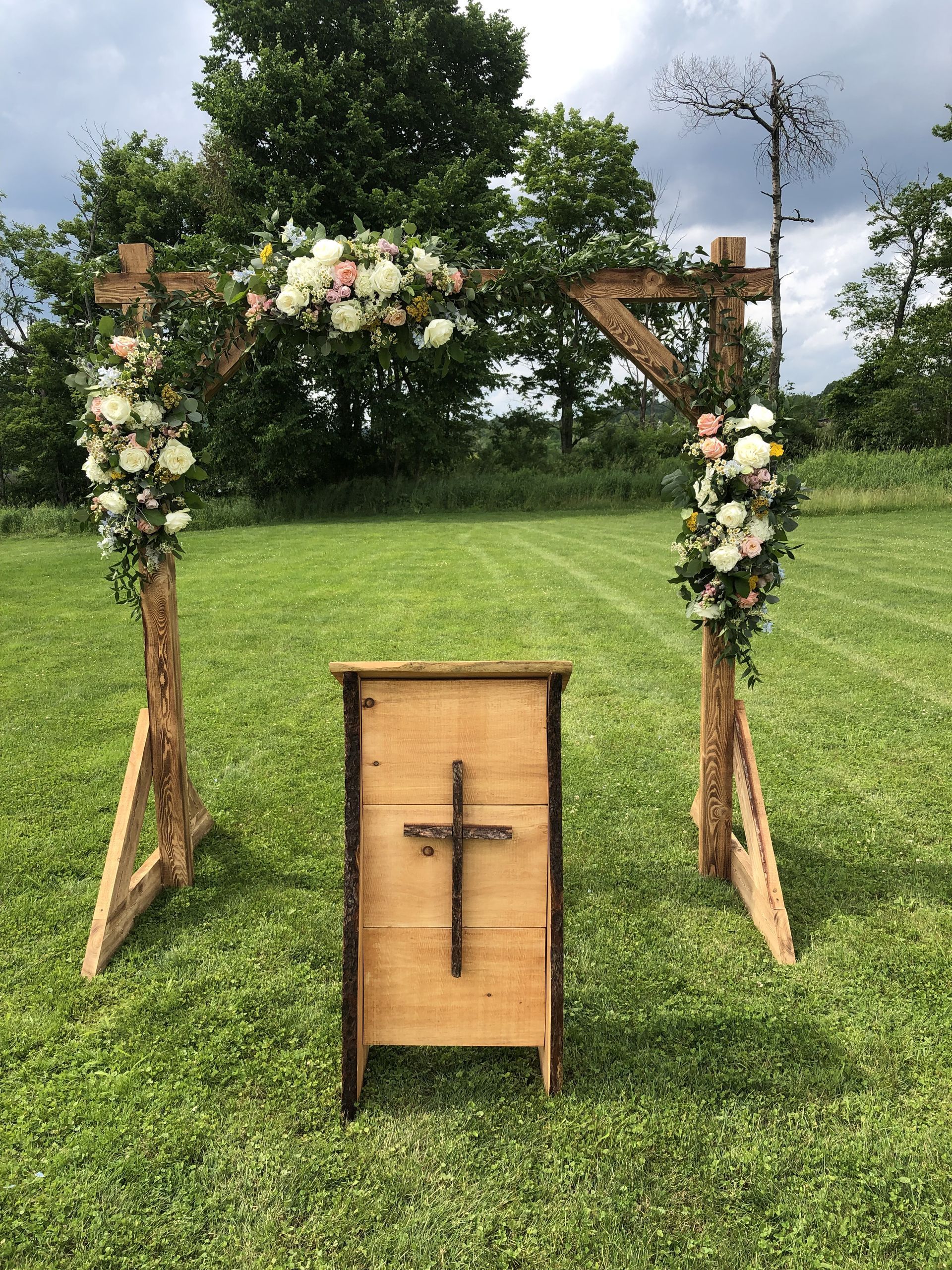 Wooden wedding arch decorated with flowers on a grassy field, with a wooden podium and cross.