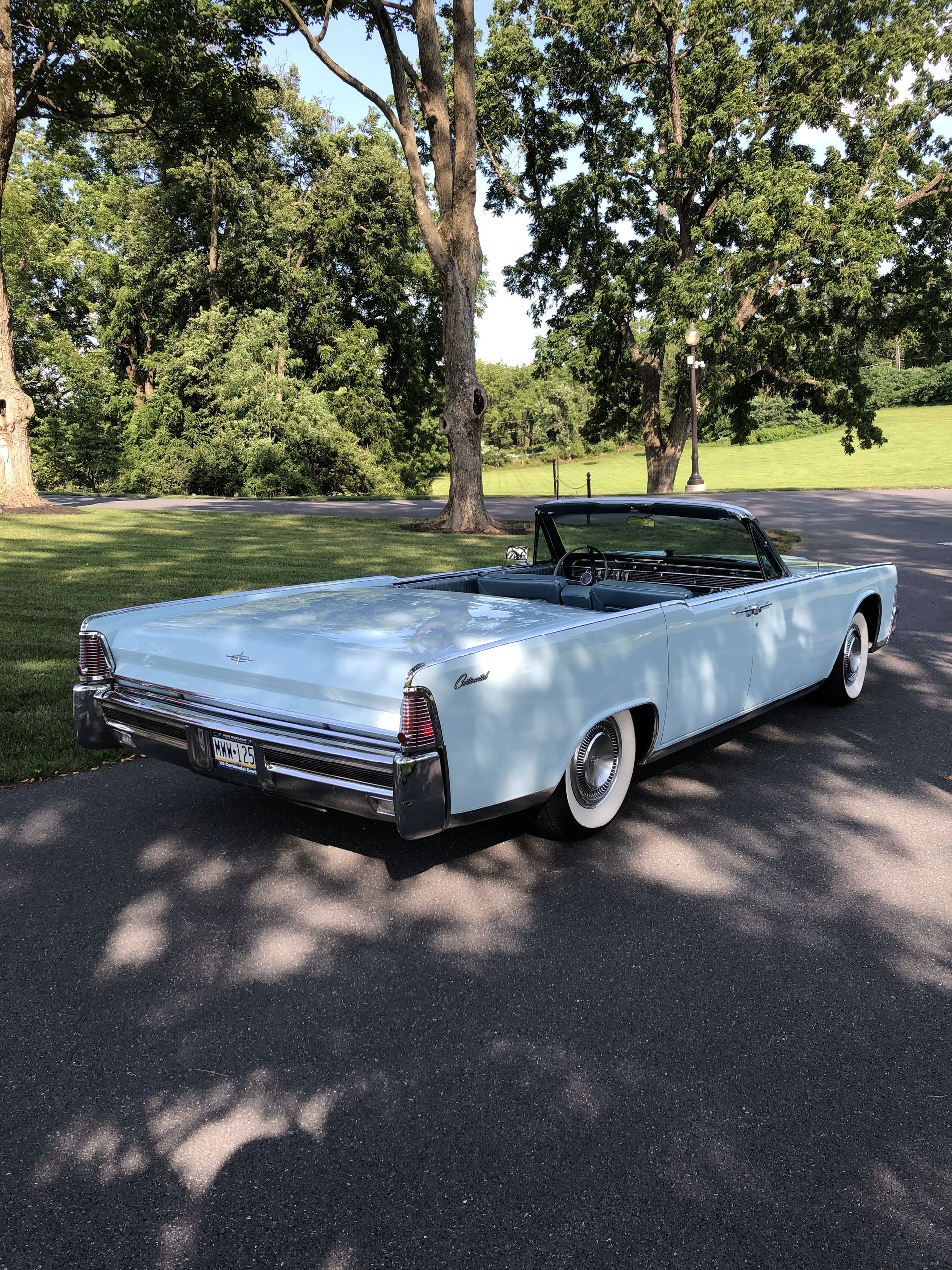 Light blue classic Lincoln Continental convertible parked on a paved road, trees in the background.