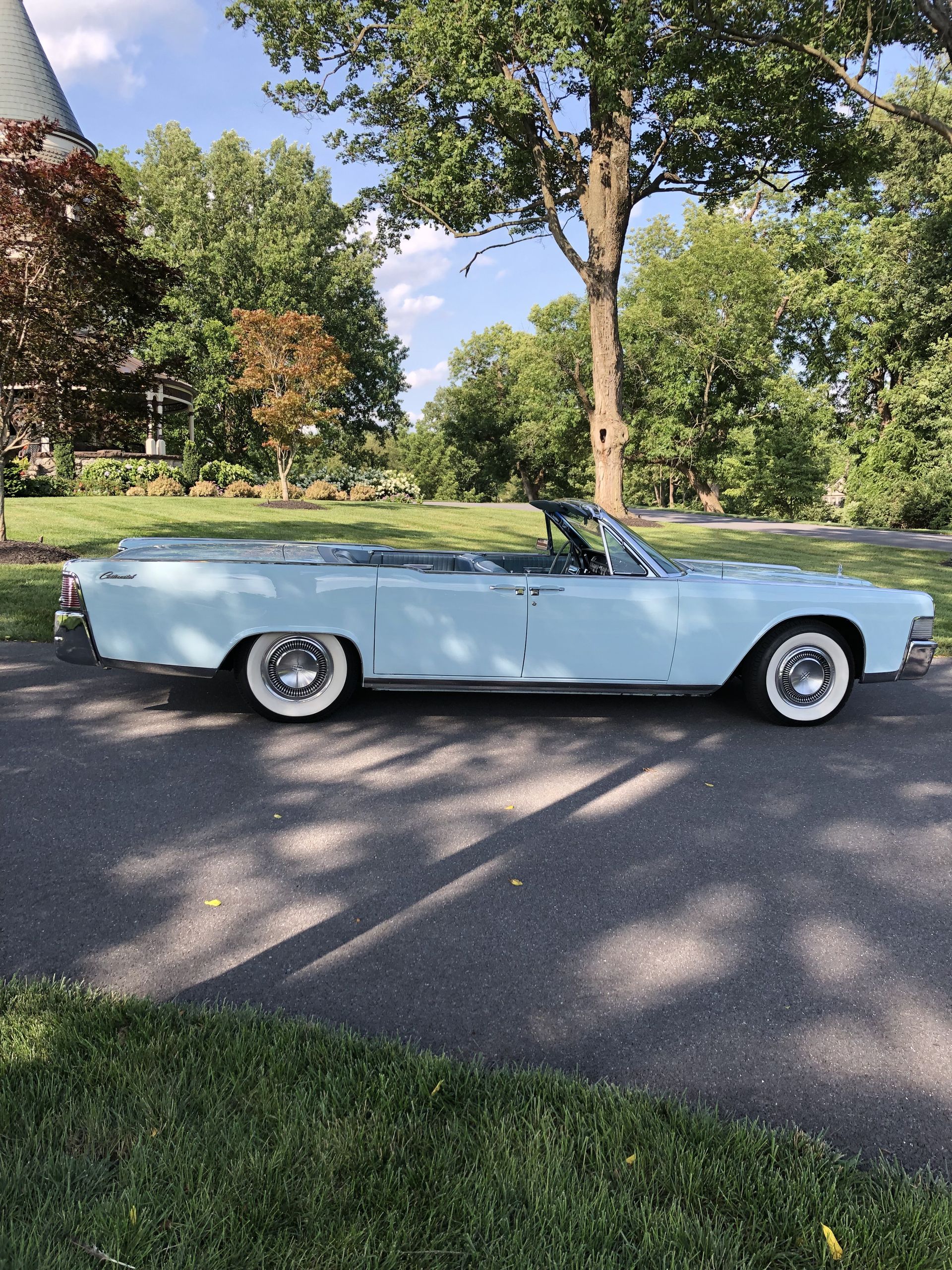Light blue convertible car with white wall tires parked on a driveway with a tree-lined background.