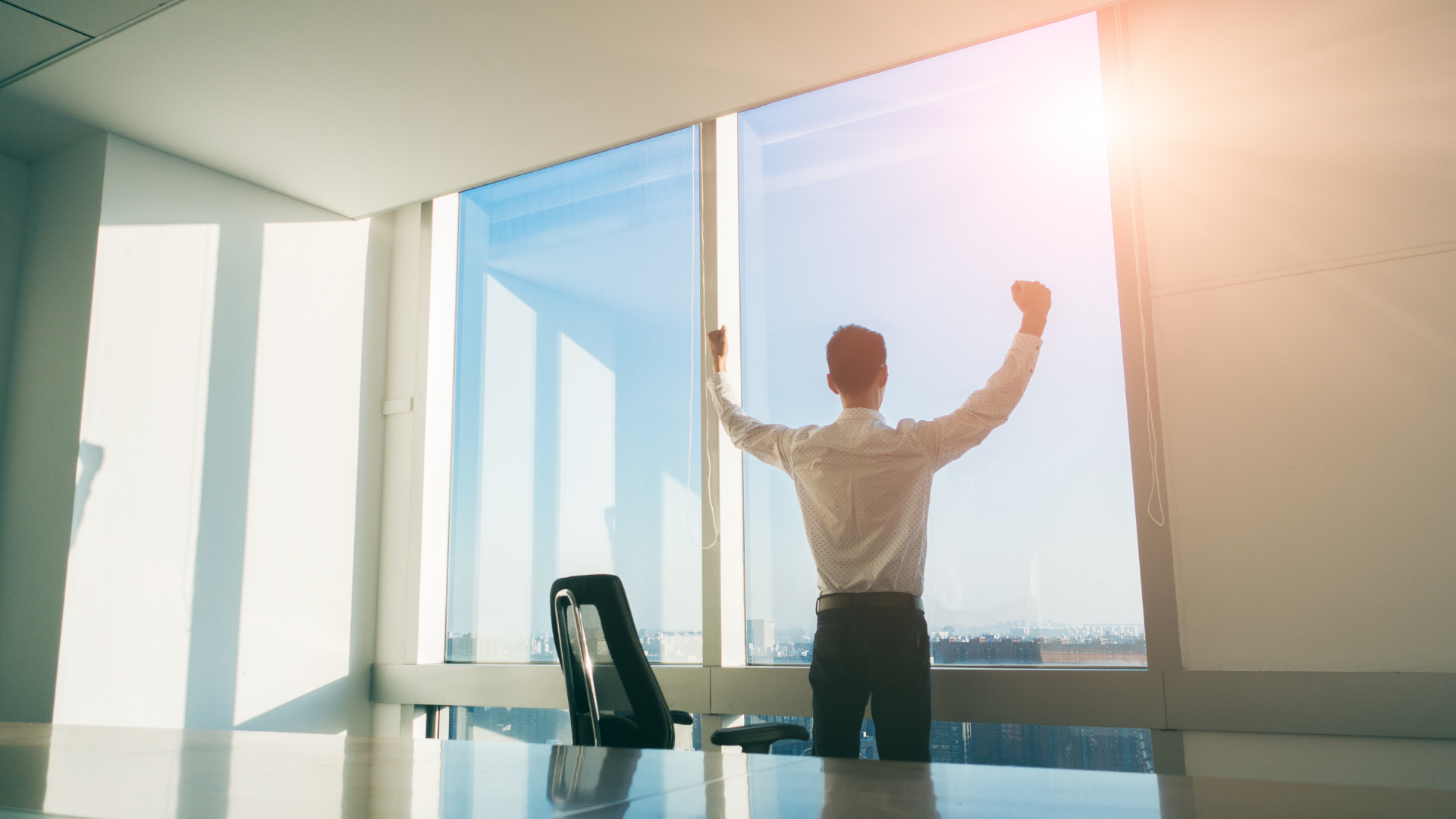 Person stretching with raised arms in a sunlit office conference room with floor-to-ceiling windows.