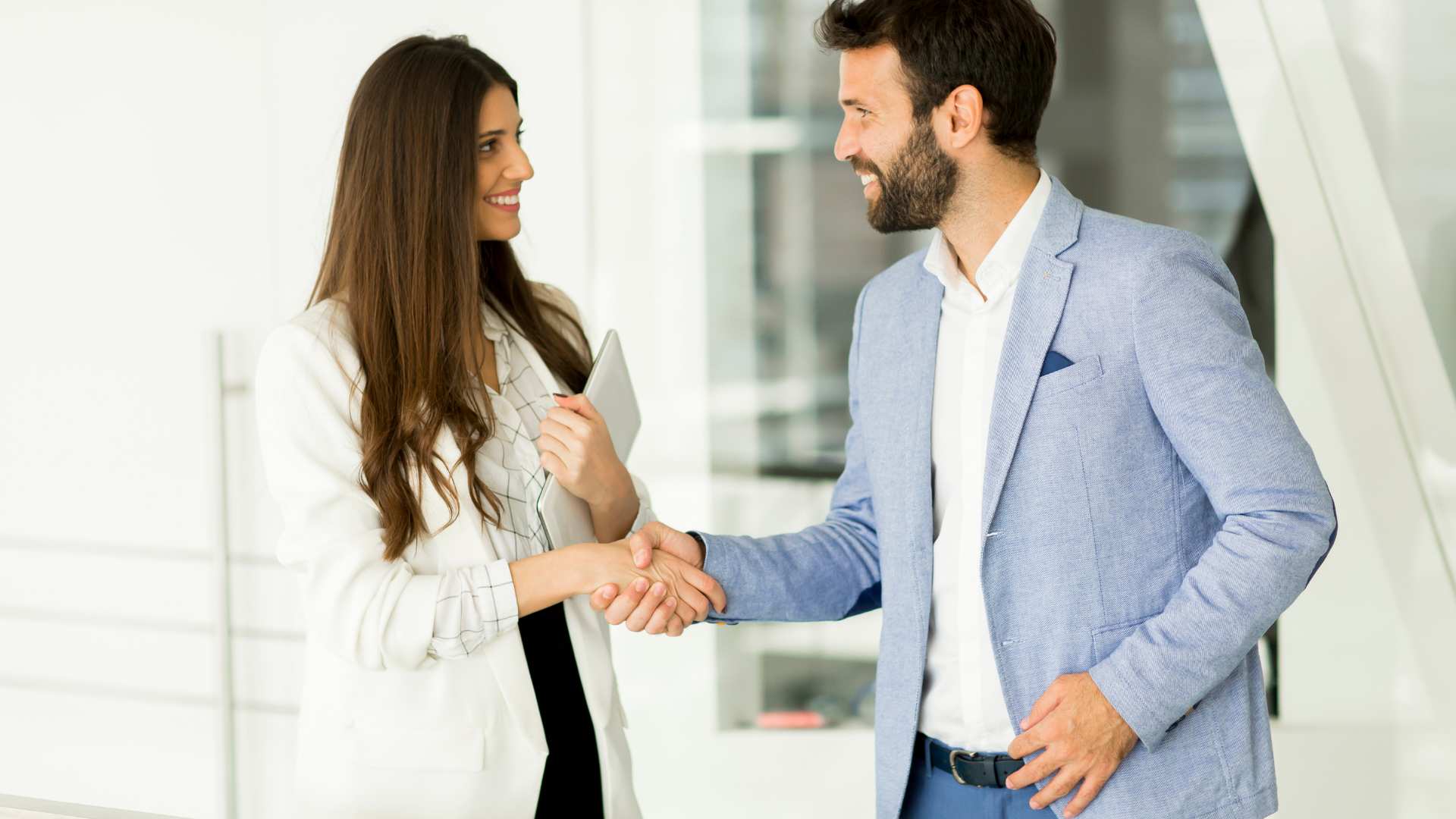 Two colleagues shaking hands in a bright office, smiling and facing each other.