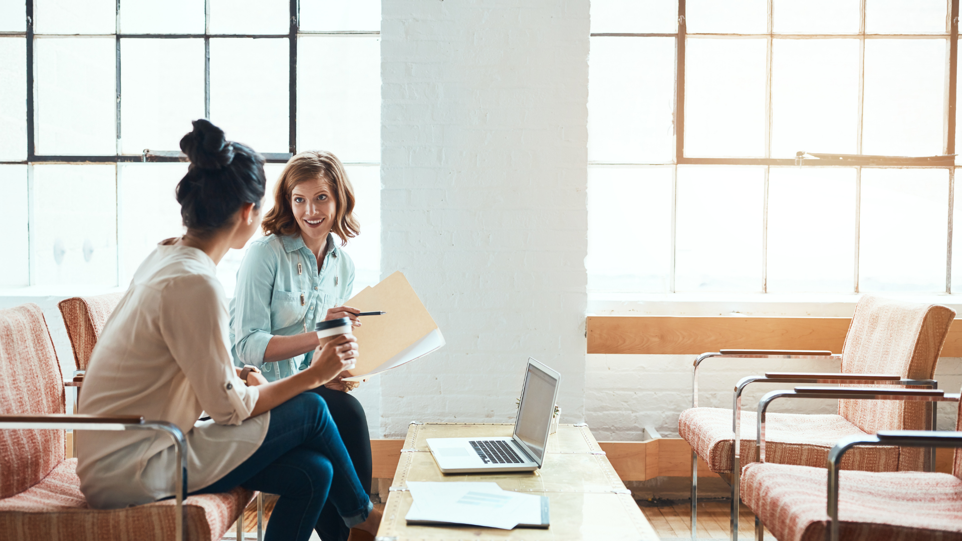 Two women in a bright office space discuss documents, one holding a coffee cup. A laptop sits on a table between them.