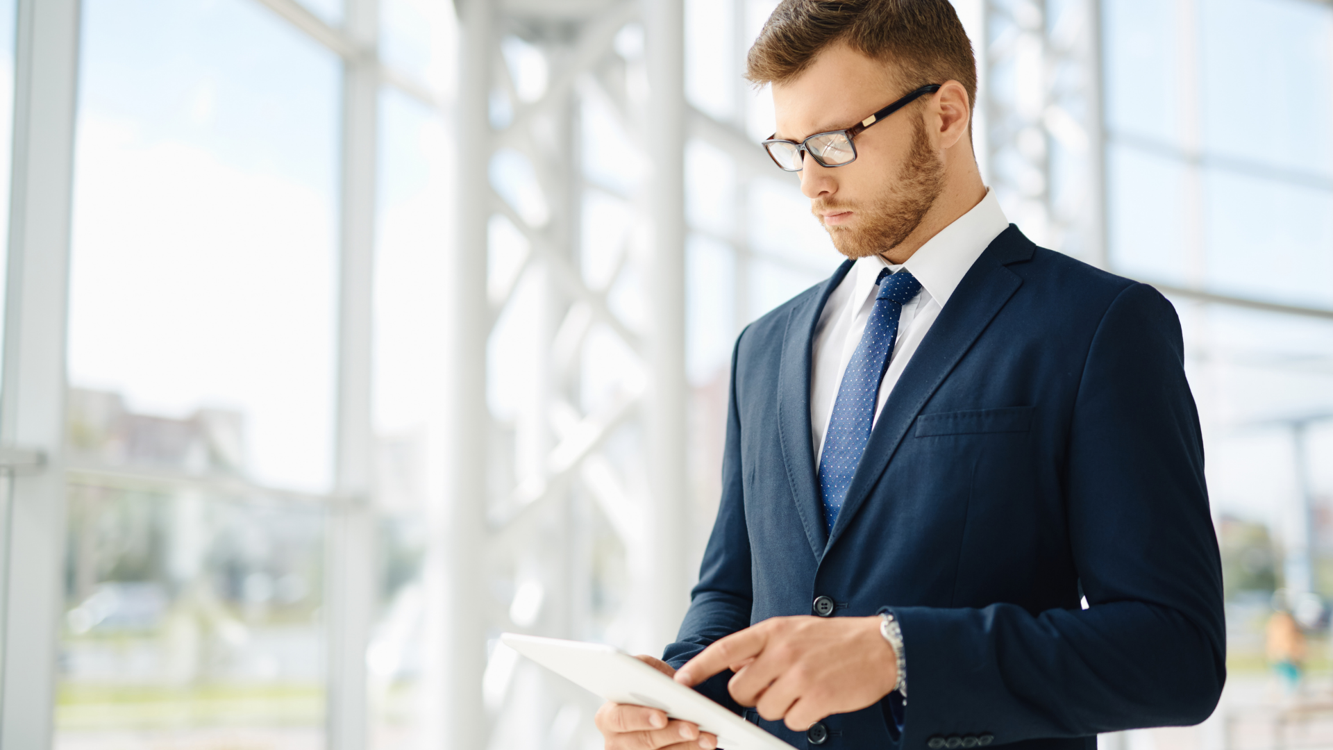 Businessman in a navy suit and glasses using a tablet near a window.
