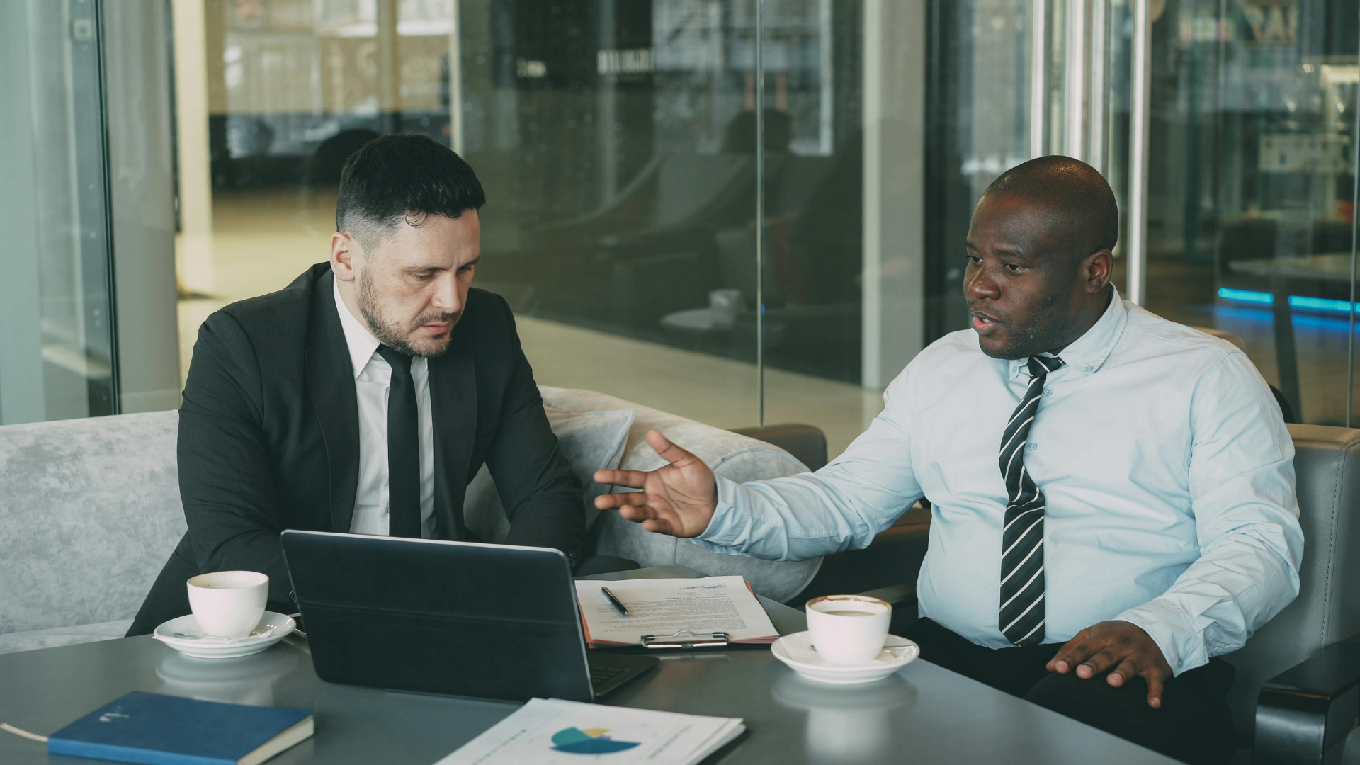 Two people in business attire discussing over laptops at a conference table in a glass-walled office