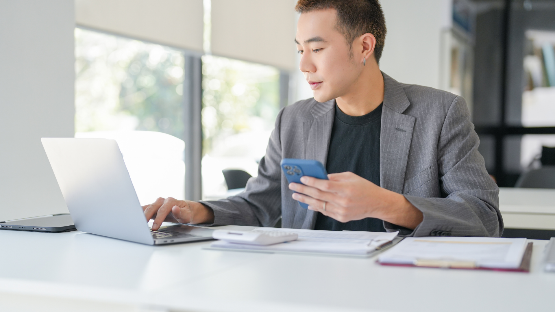 Person working on a laptop while holding a phone at a bright desk in an office