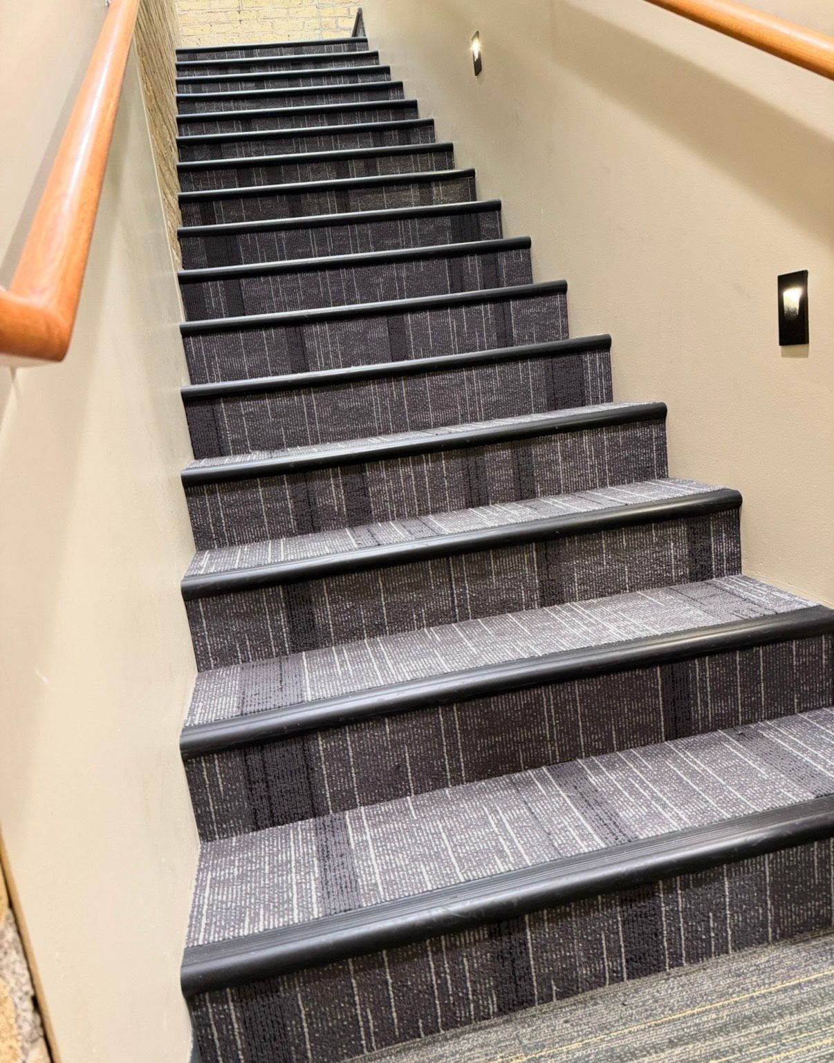 Staircase with gray carpeted steps, dark trim, and wooden handrails on a beige wall.