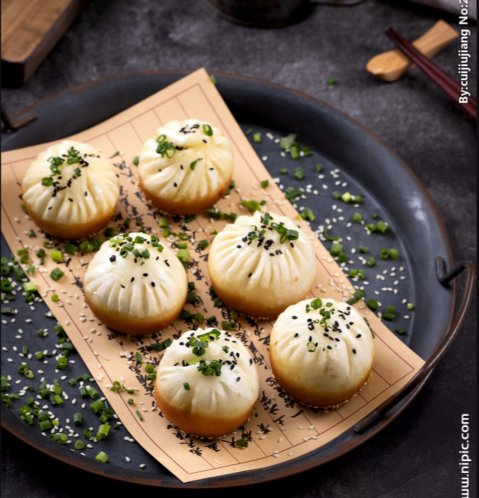 Steamed dumplings on a bamboo tray on a dark plate, topped with sesame seeds and chopped herbs.