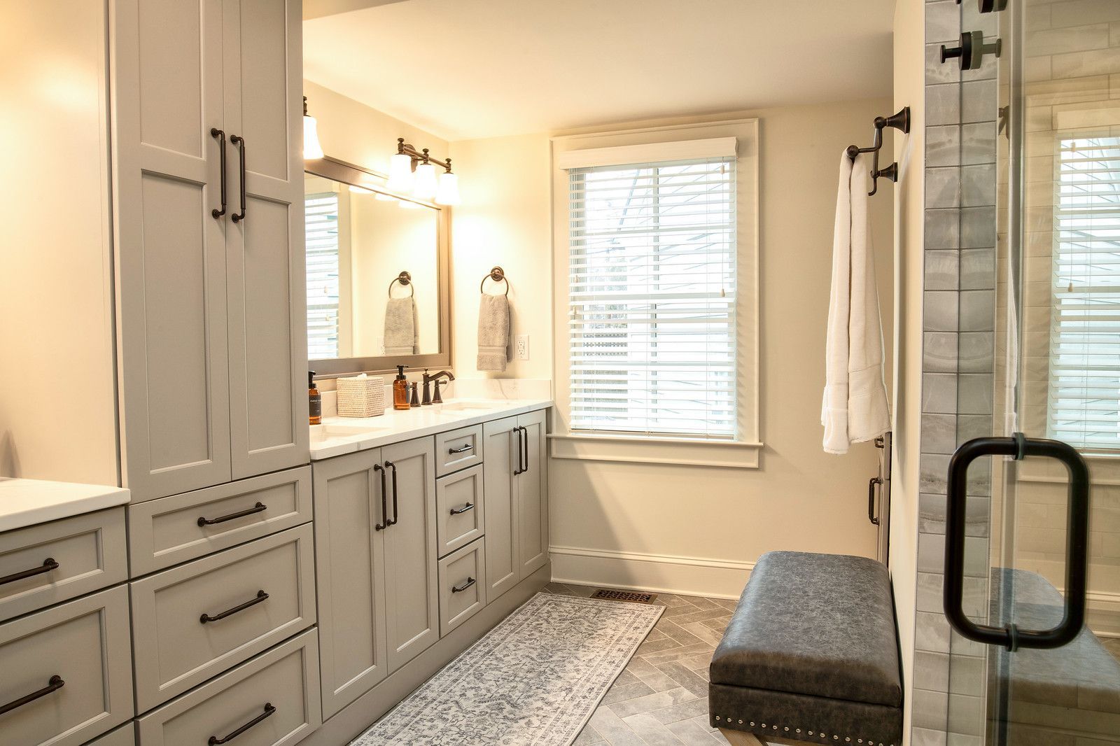 Bright modern bathroom with gray cabinets, double vanity, shower, window, and bench.
