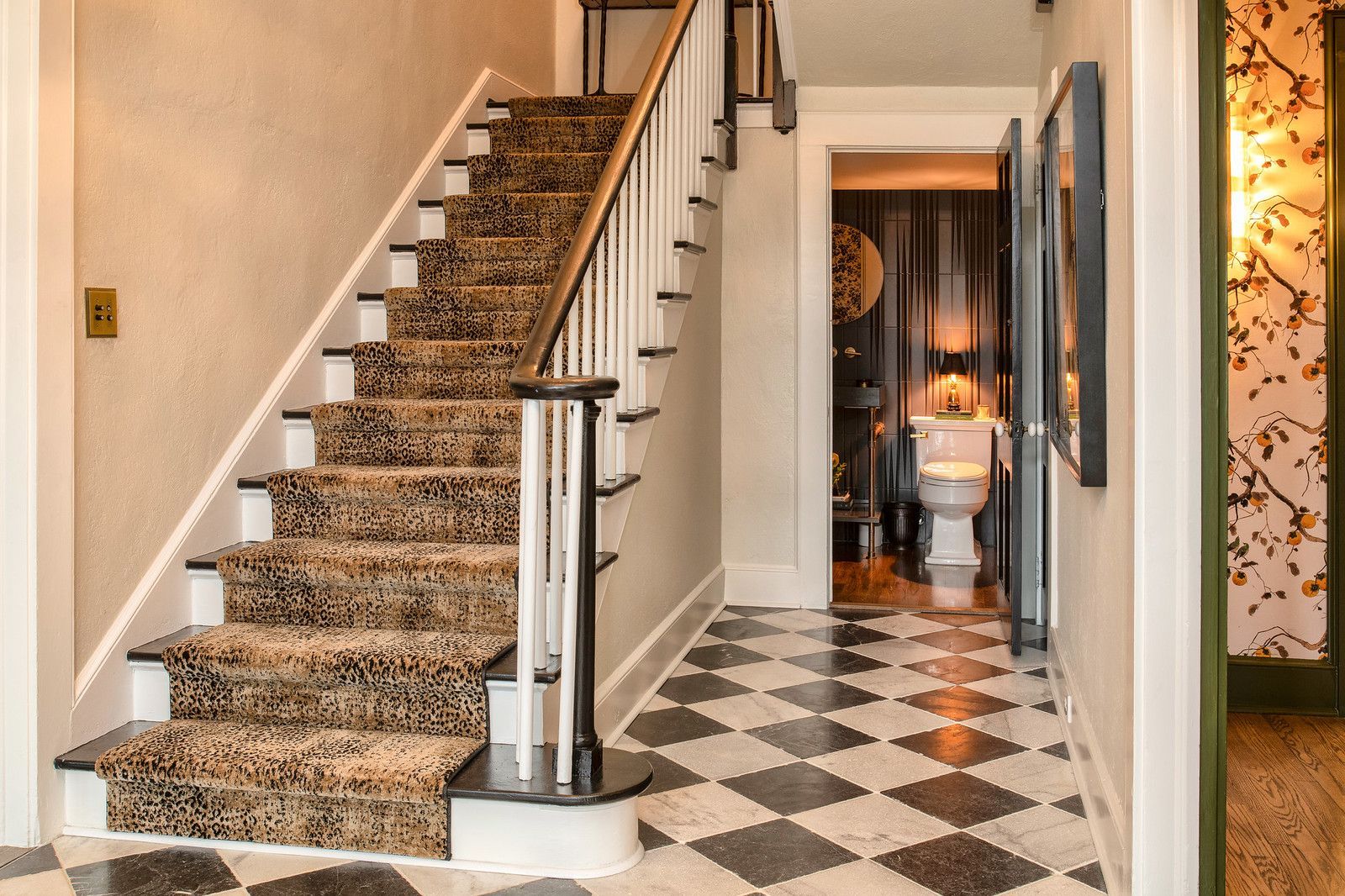 Hallway with staircase, patterned carpet runner, and checkered tile floor leading to a bathroom.
