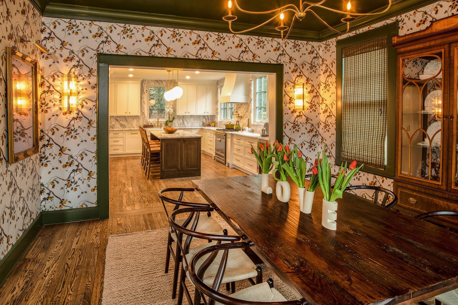 Cozy dining room with a long wooden table, bar stools, floral wallpaper, and kitchen visible through archway