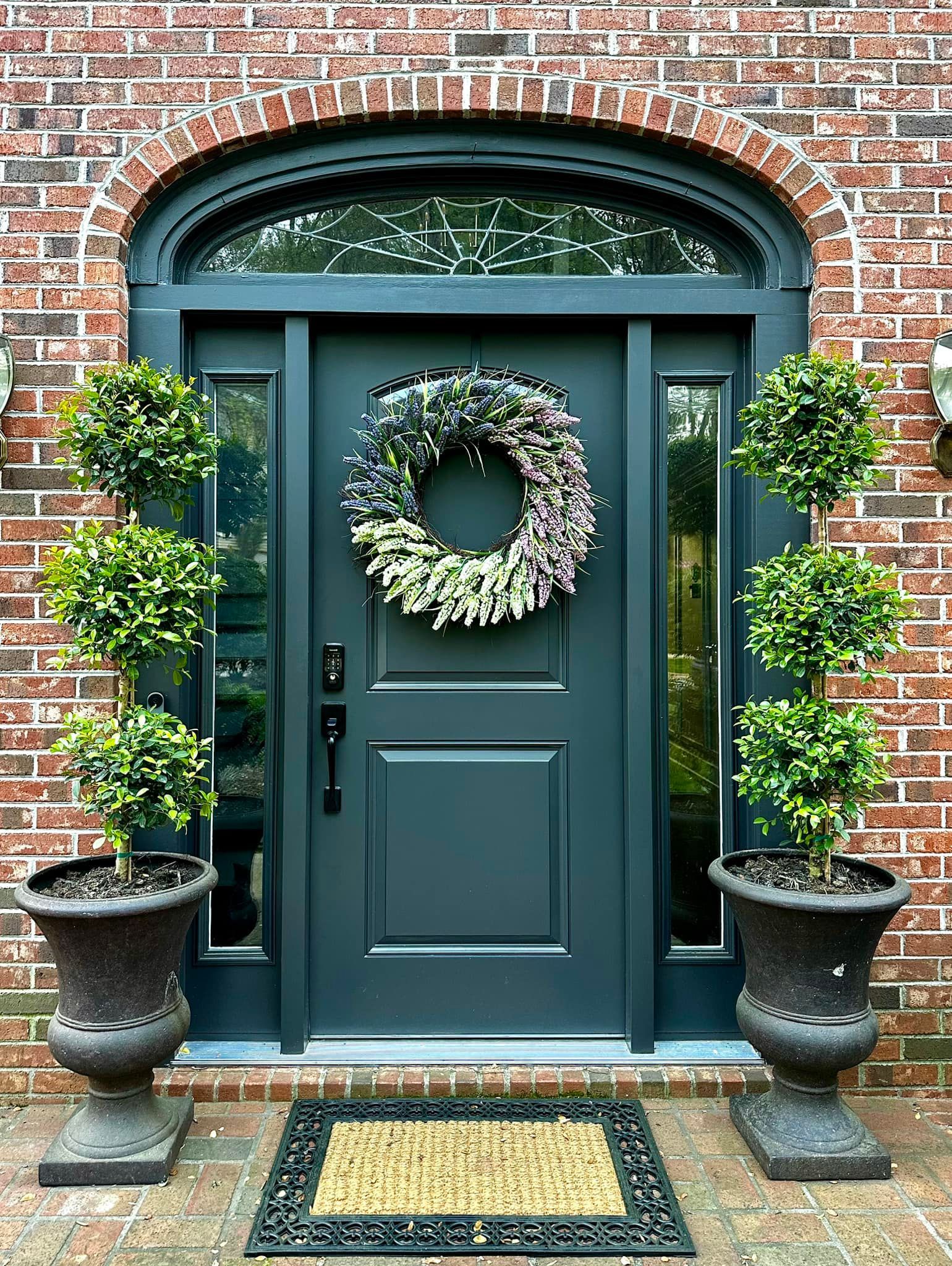 Dark teal front door with wreath, flanked by potted topiary trees under a brick archway.
