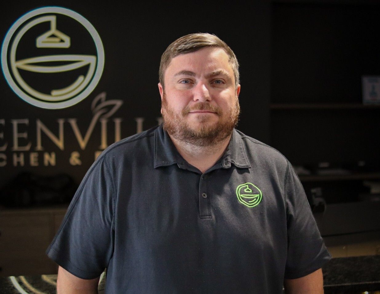 Man standing in front of a business sign, wearing a dark polo shirt with a green logo.