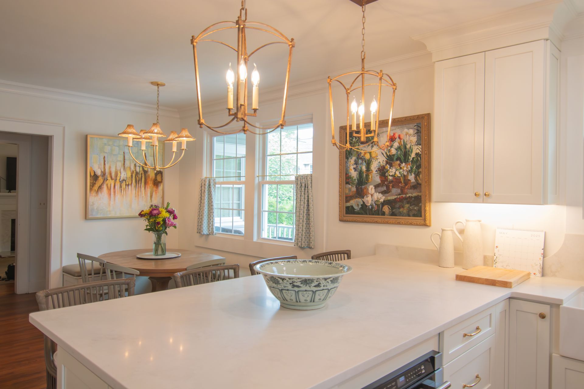 Bright white kitchen with marble island, gold chandeliers, dining table, and floral wall art