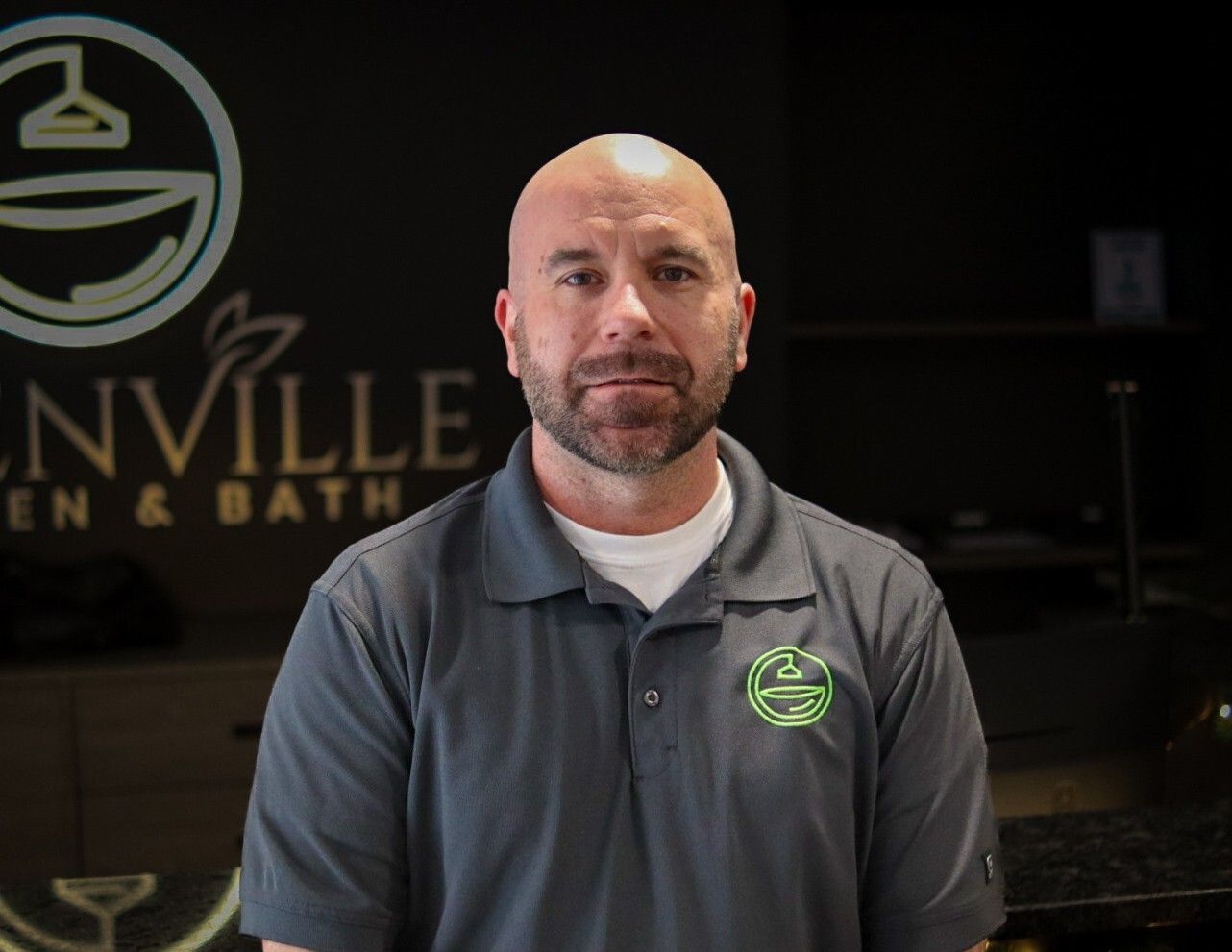 Man in a gray polo shirt standing in a showroom with a logo sign in the background