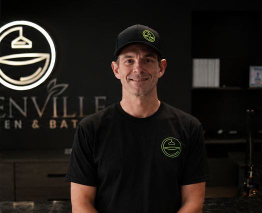 Smiling man in black shirt and cap standing in a dark gym lobby with a neon logo sign behind him