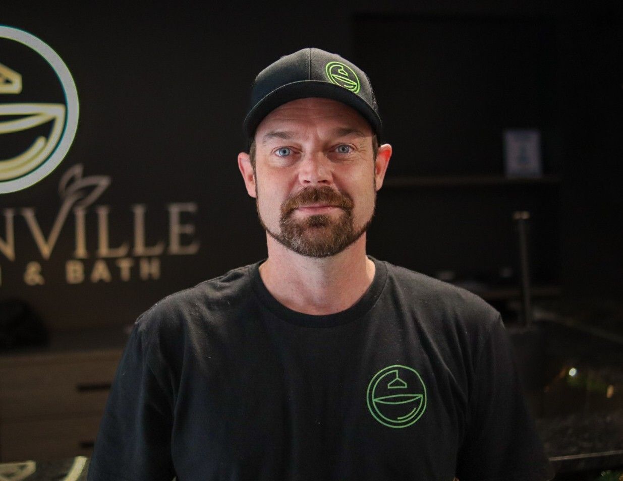 Man in black shirt and cap standing indoors in front of a business logo sign