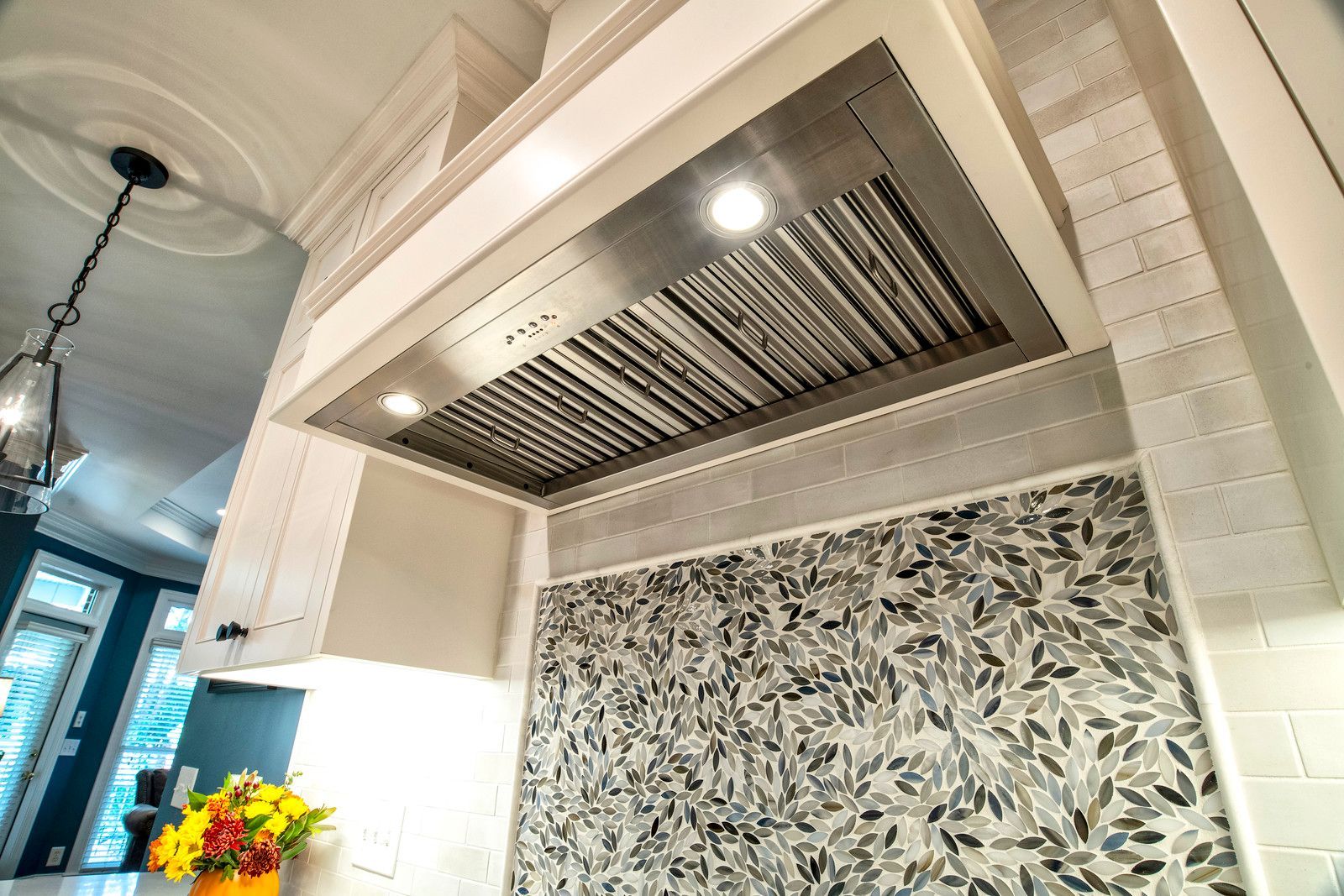 Modern kitchen range hood with stainless vent, patterned backsplash, and recessed light above counter