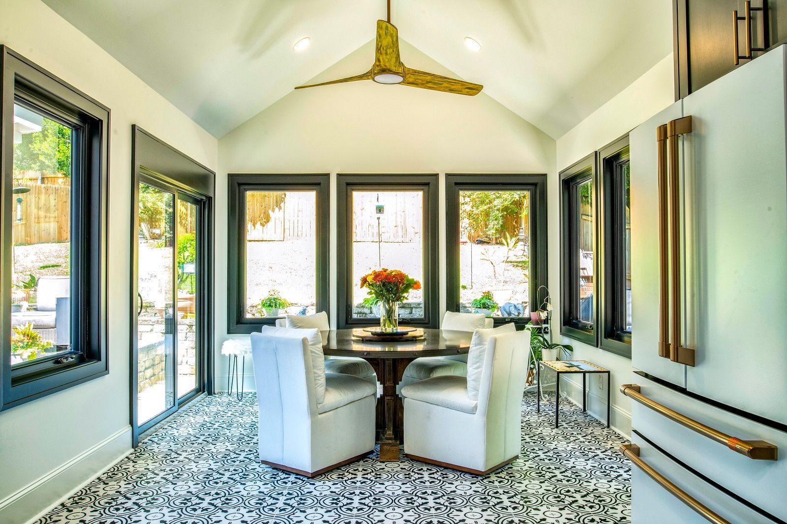 Bright modern sunroom dining area with white chairs, patterned floor, and large windows overlooking greenery