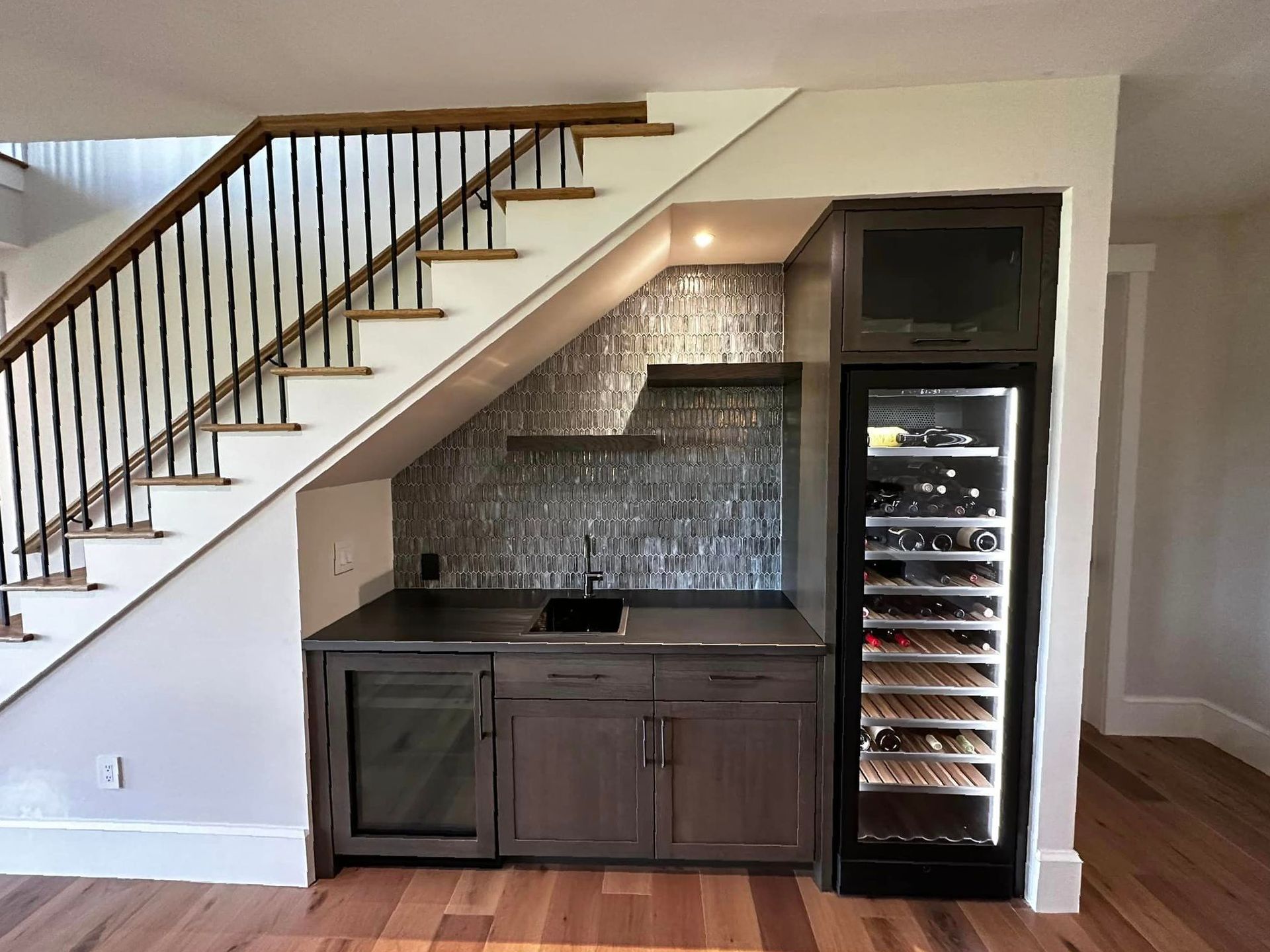 Under-stair wet bar with dark cabinets, tile backsplash, and wine fridge in a modern home interior