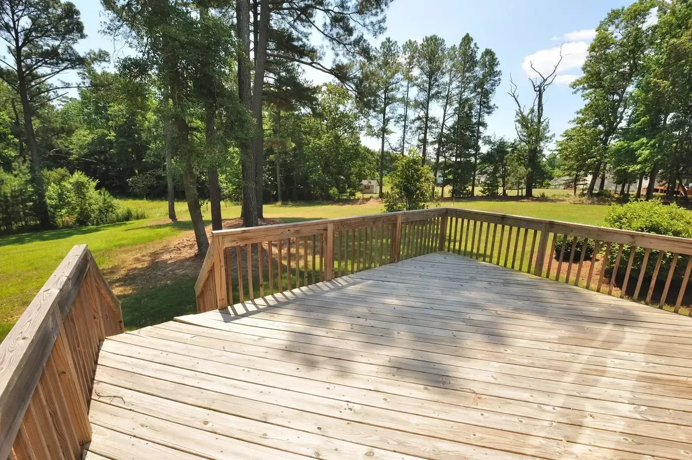 Wooden deck overlooking a sunny grassy yard with tall trees and a blue sky