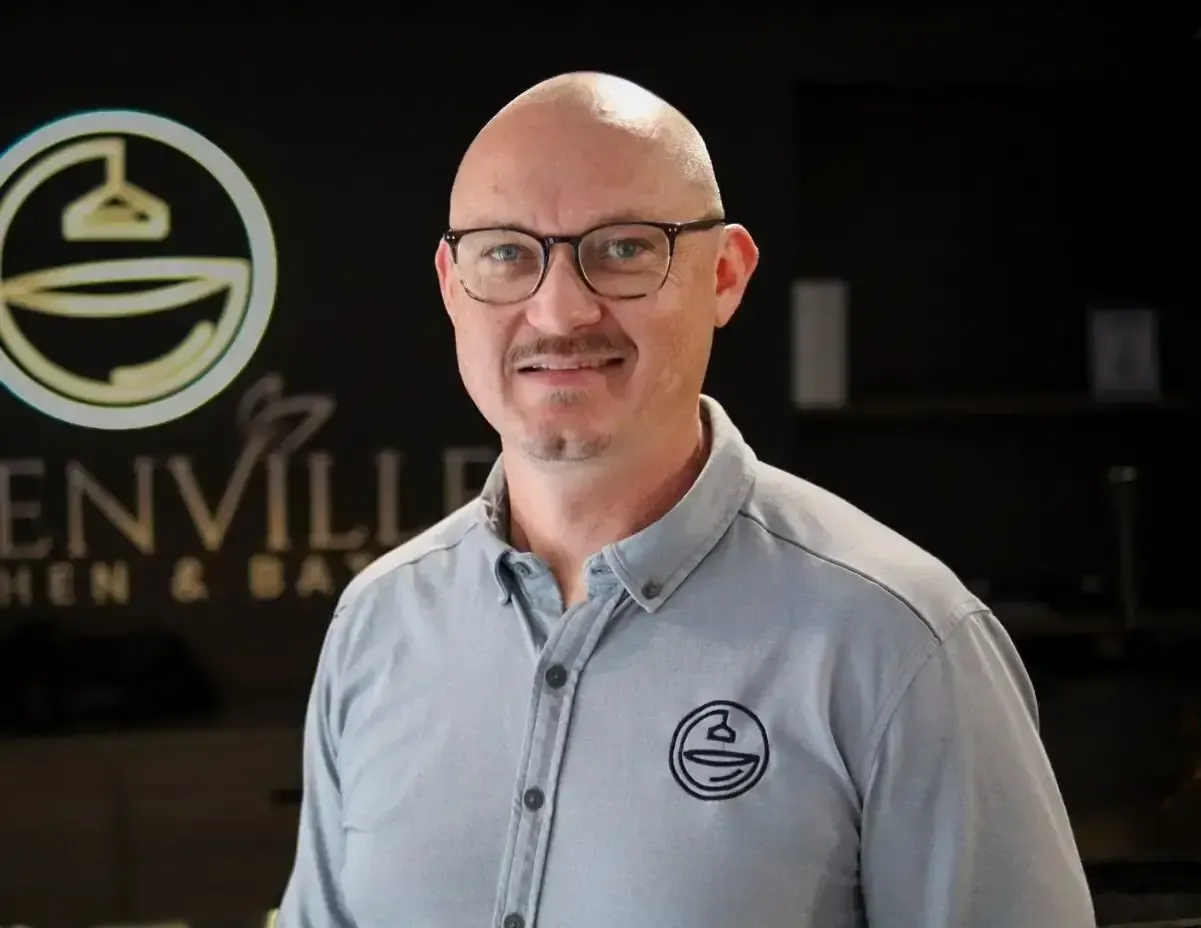 Smiling man in glasses wearing a gray polo, standing indoors near a business logo backdrop.
