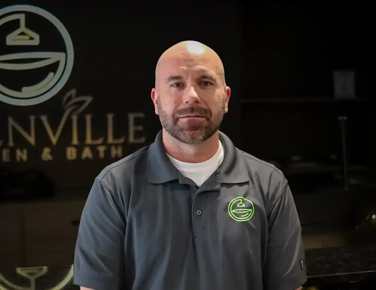 Man in gray polo stands in front of a dark showroom sign reading “...VILLE Bath & Bath”