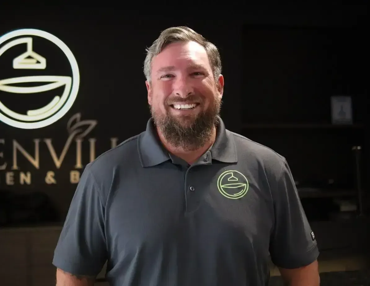 Smiling man in a gray polo stands in front of a company logo and storefront sign.