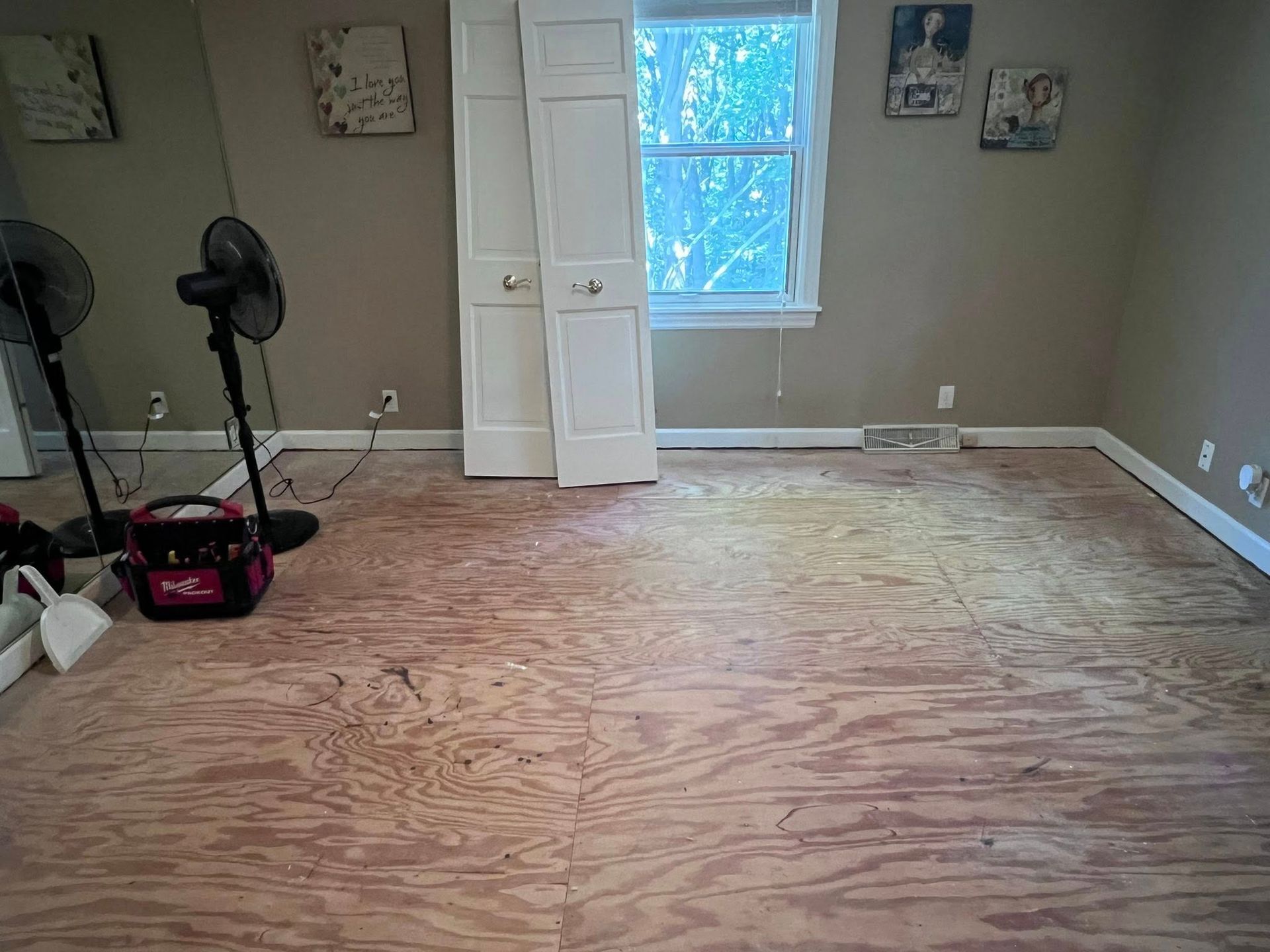 Empty room with plywood subfloor, window, fan, and a few tools along the wall.