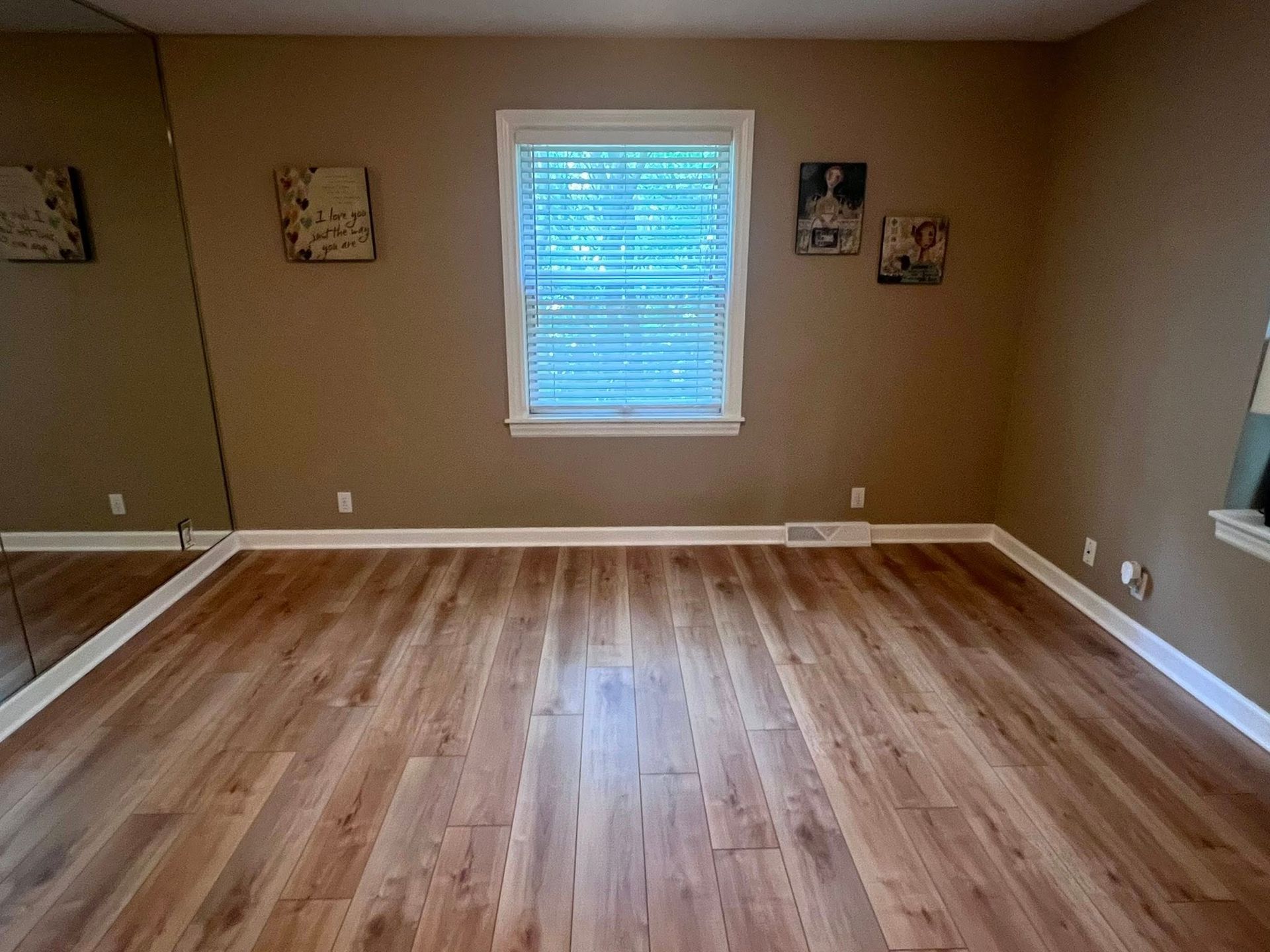 Empty beige room with hardwood floors and a central window with blinds.