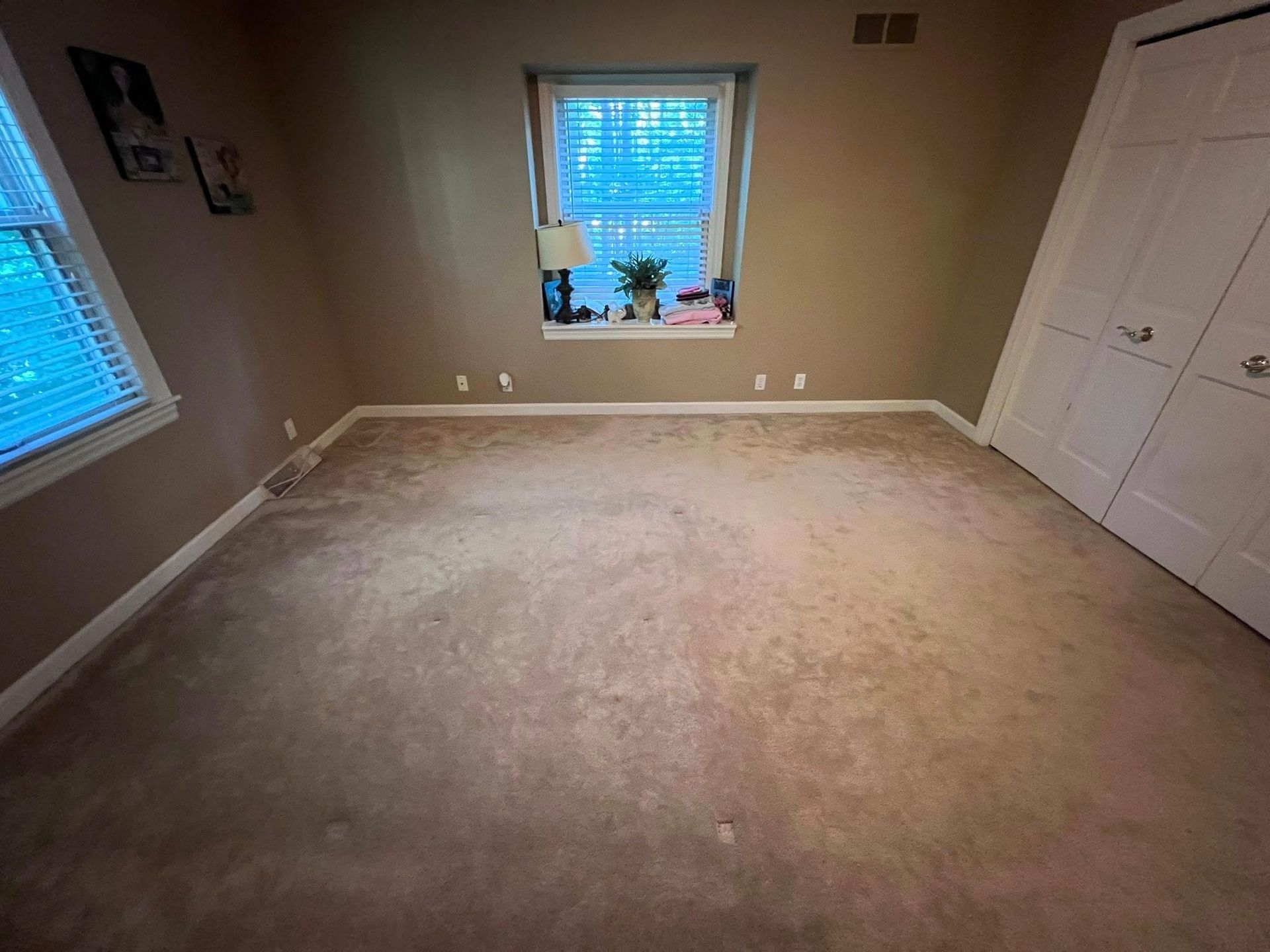 Empty beige bedroom with carpet, two windows with blinds, and white double closet doors