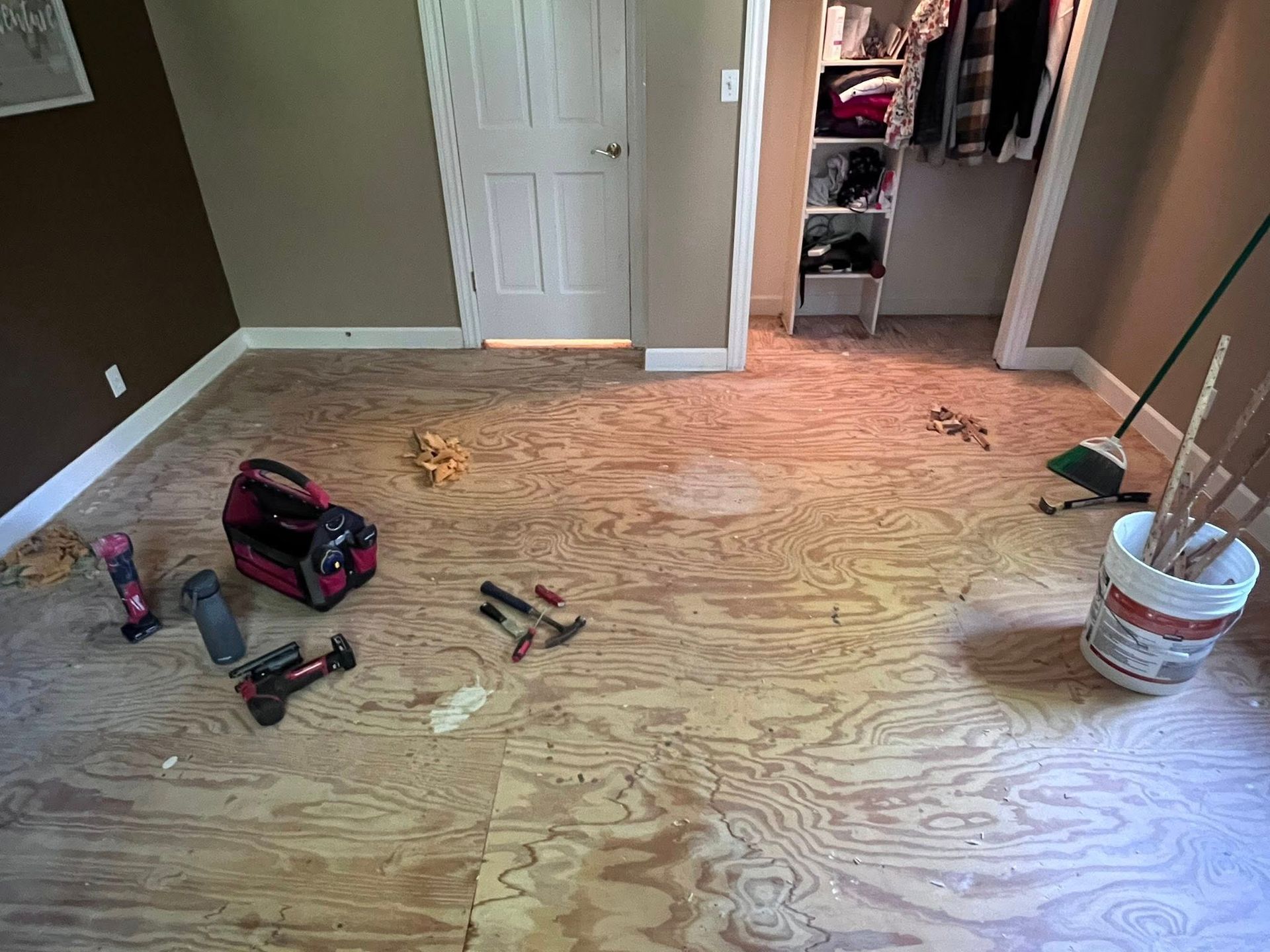 Partially renovated room with plywood subfloor, tools, and a bucket of supplies