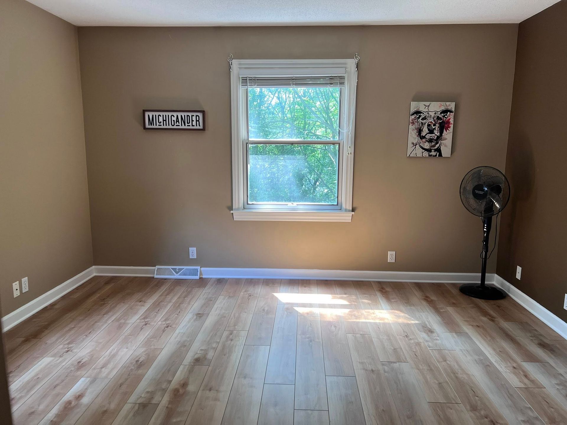 Empty beige room with hardwood floors, a centered window, wall art, and a standing fan in the corner