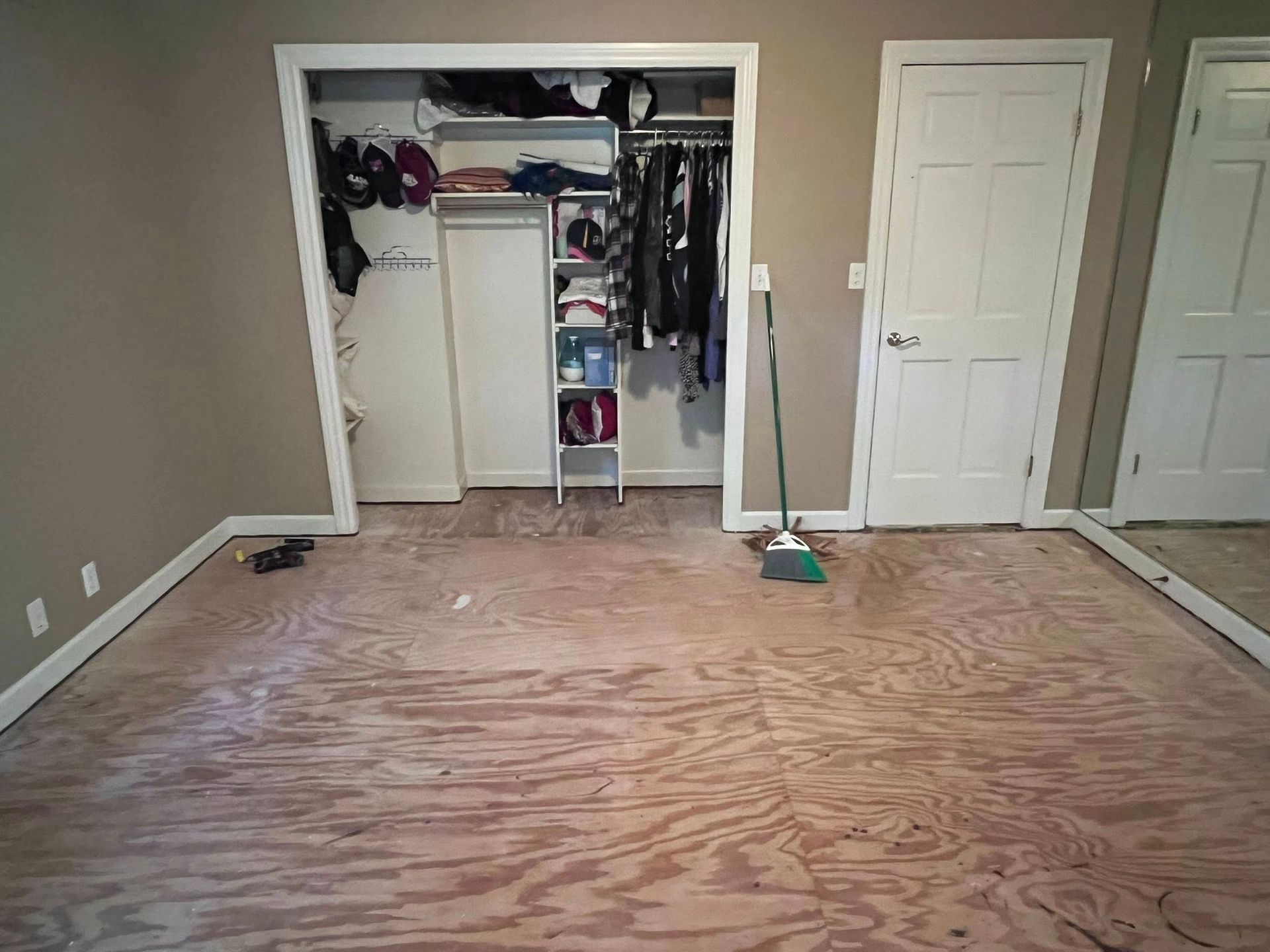 Empty bedroom with bare plywood floor, open closet, and white door against beige walls.