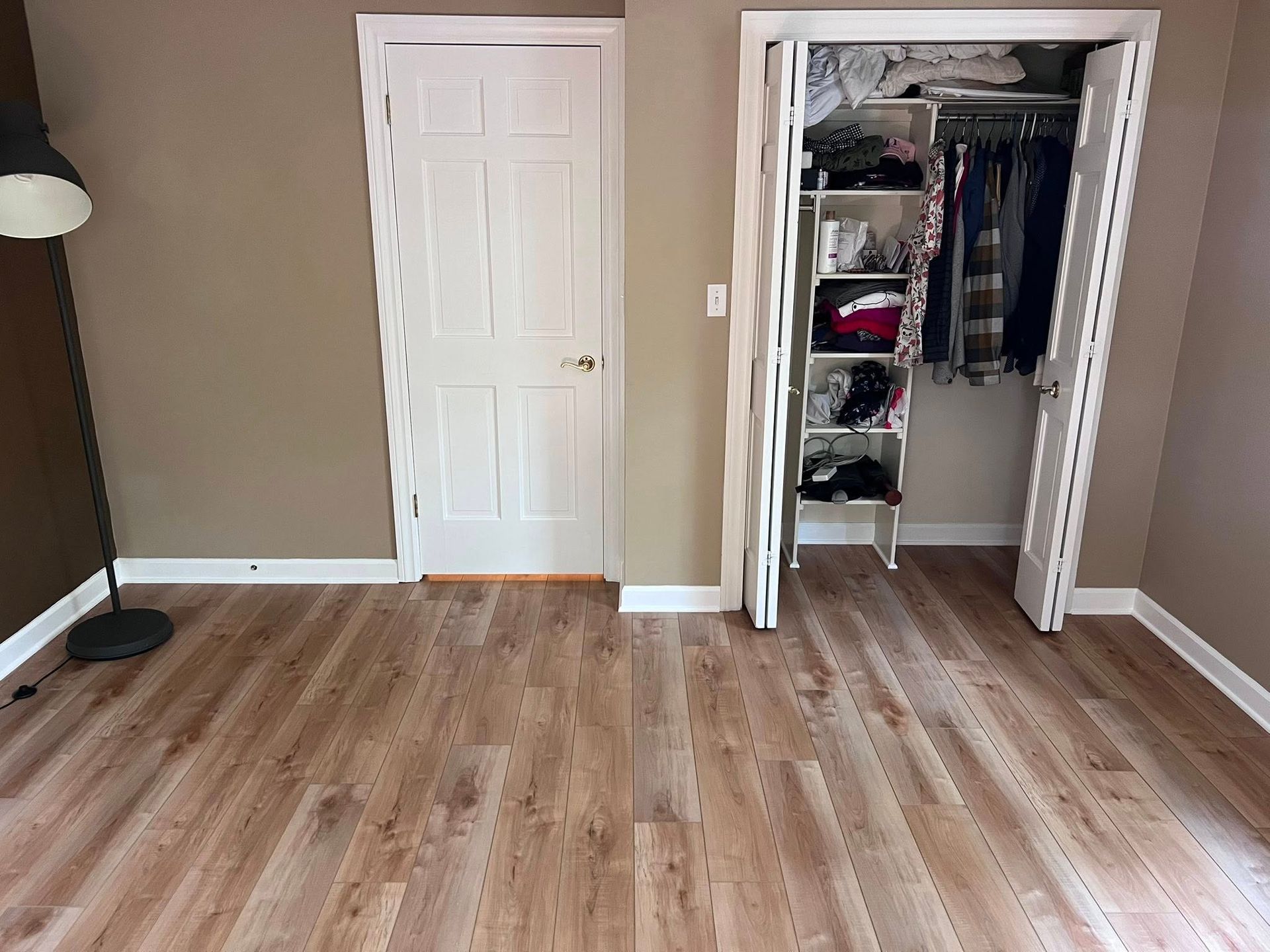Empty bedroom with wood floor, beige walls, floor lamp, white closet door, and open closet with hanging clothes