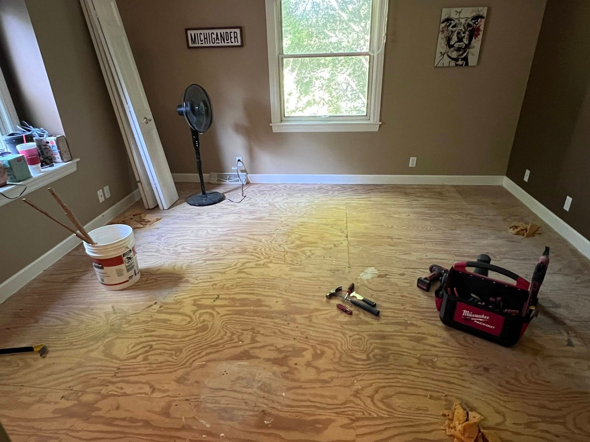 Empty room with plywood subfloor, brown walls, a window, and tools and paint supplies on the floor.