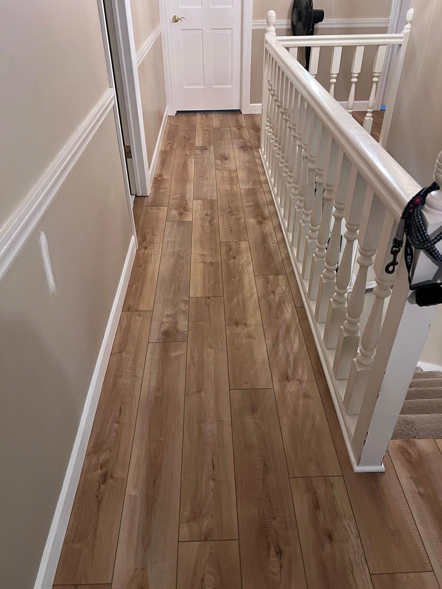 Narrow upstairs hallway with wood floor, white walls, banister, and a closed door at the end.