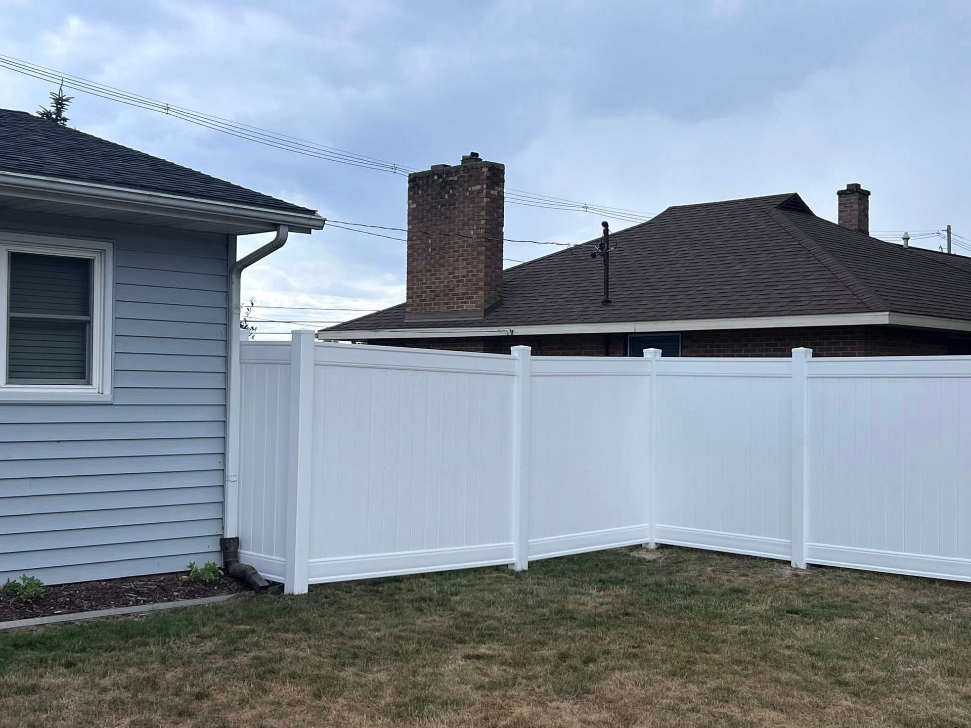 Backyard with white vinyl fence, blue house siding, and neighboring brick chimneys under a cloudy sky