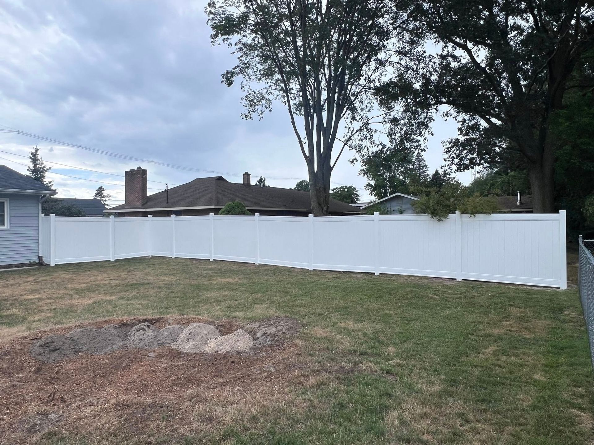 Backyard with white vinyl fence, grassy lawn, and a tree under a cloudy sky.