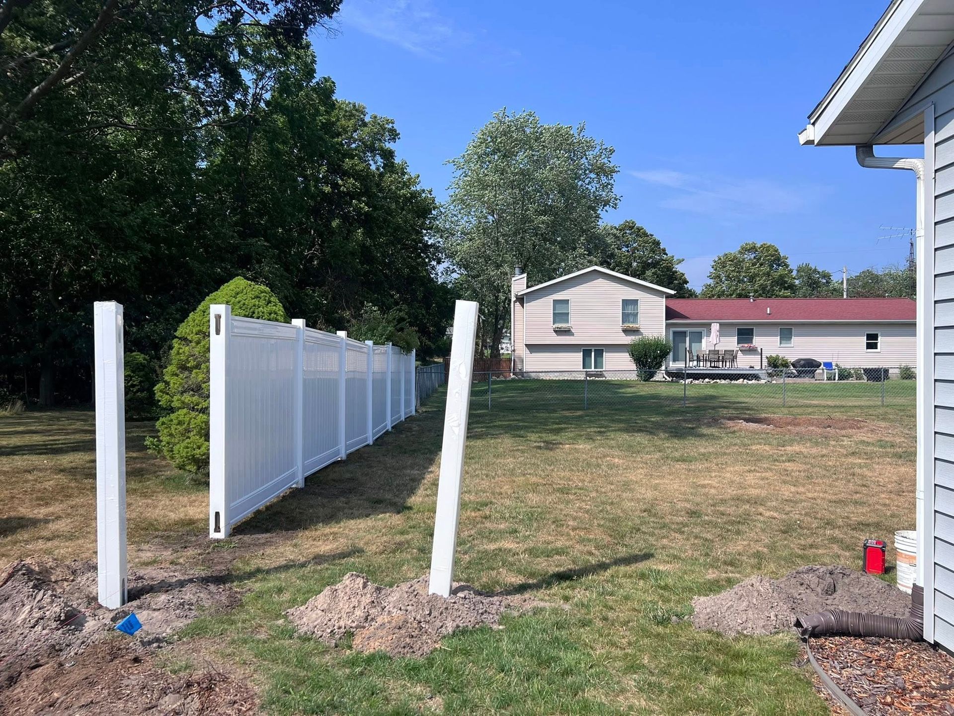 White fence posts line a yard beside a house with a red roof under a blue sky.