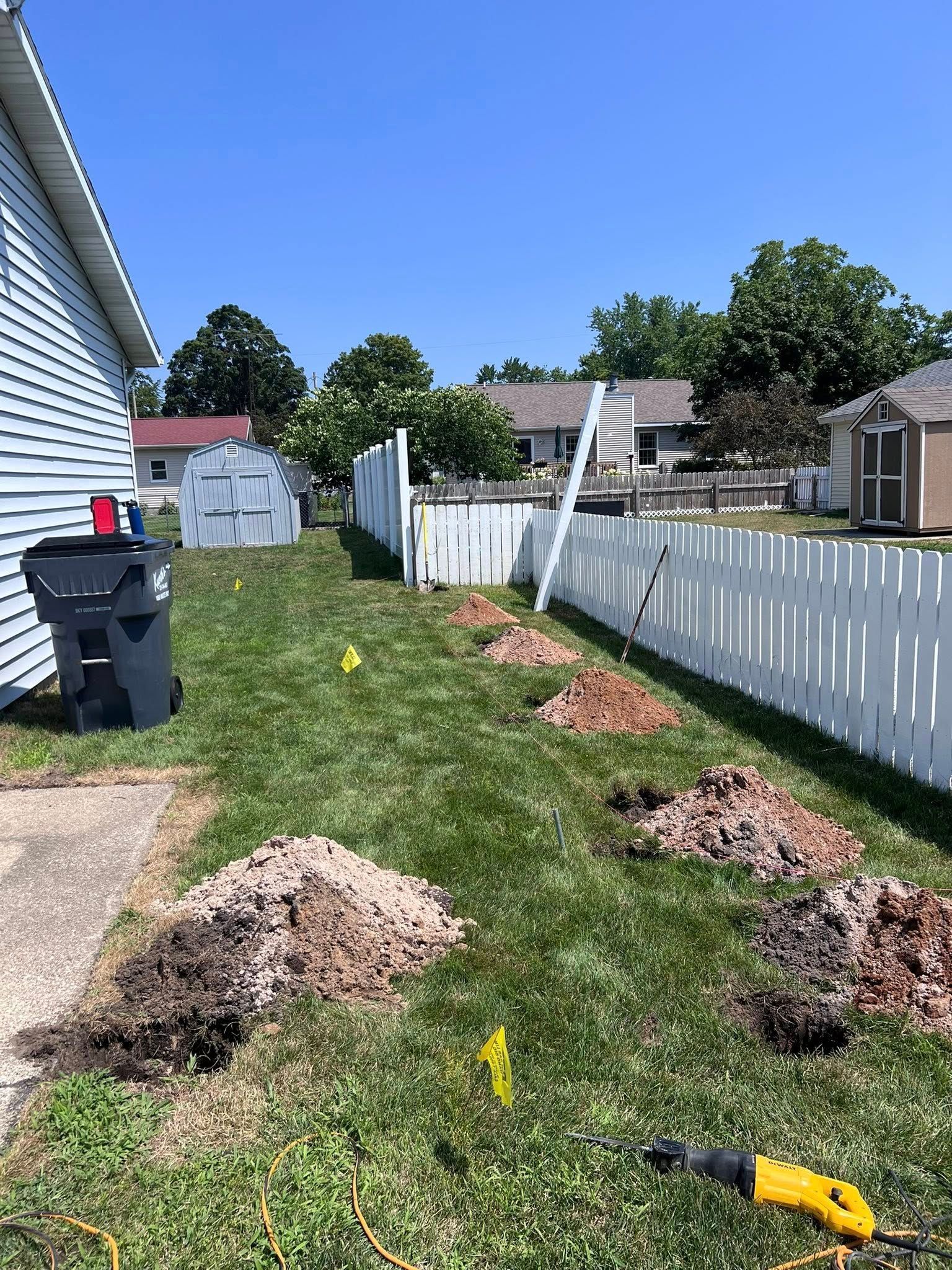 Backyard with dirt mounds, yellow flags, white fence, and a yellow power tool on grass beside a house