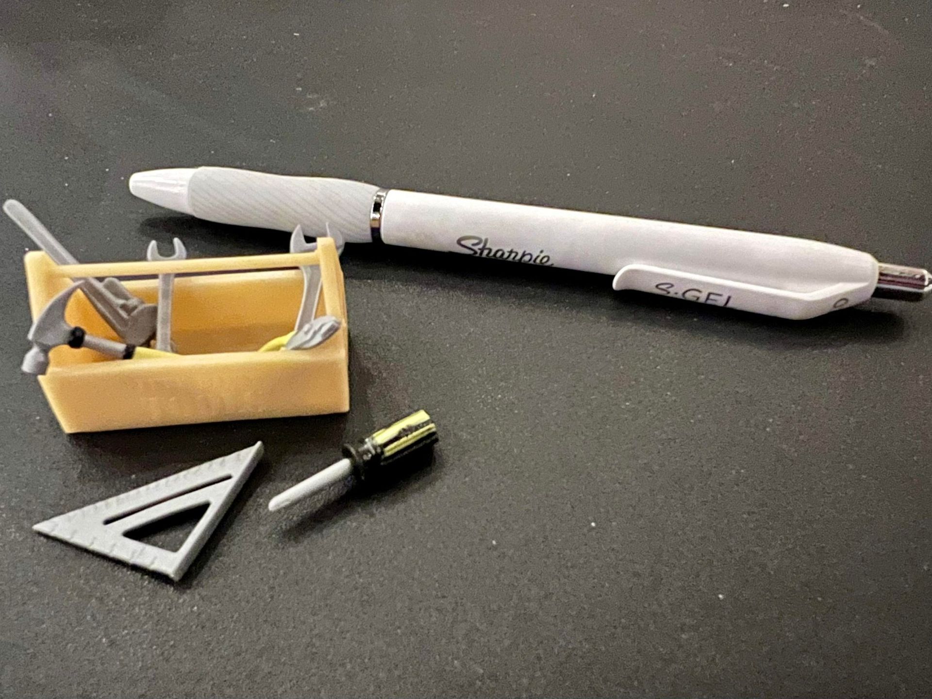 White pen tool, small yellow tray, metal triangle, and a tiny screwdriver on a dark tabletop.