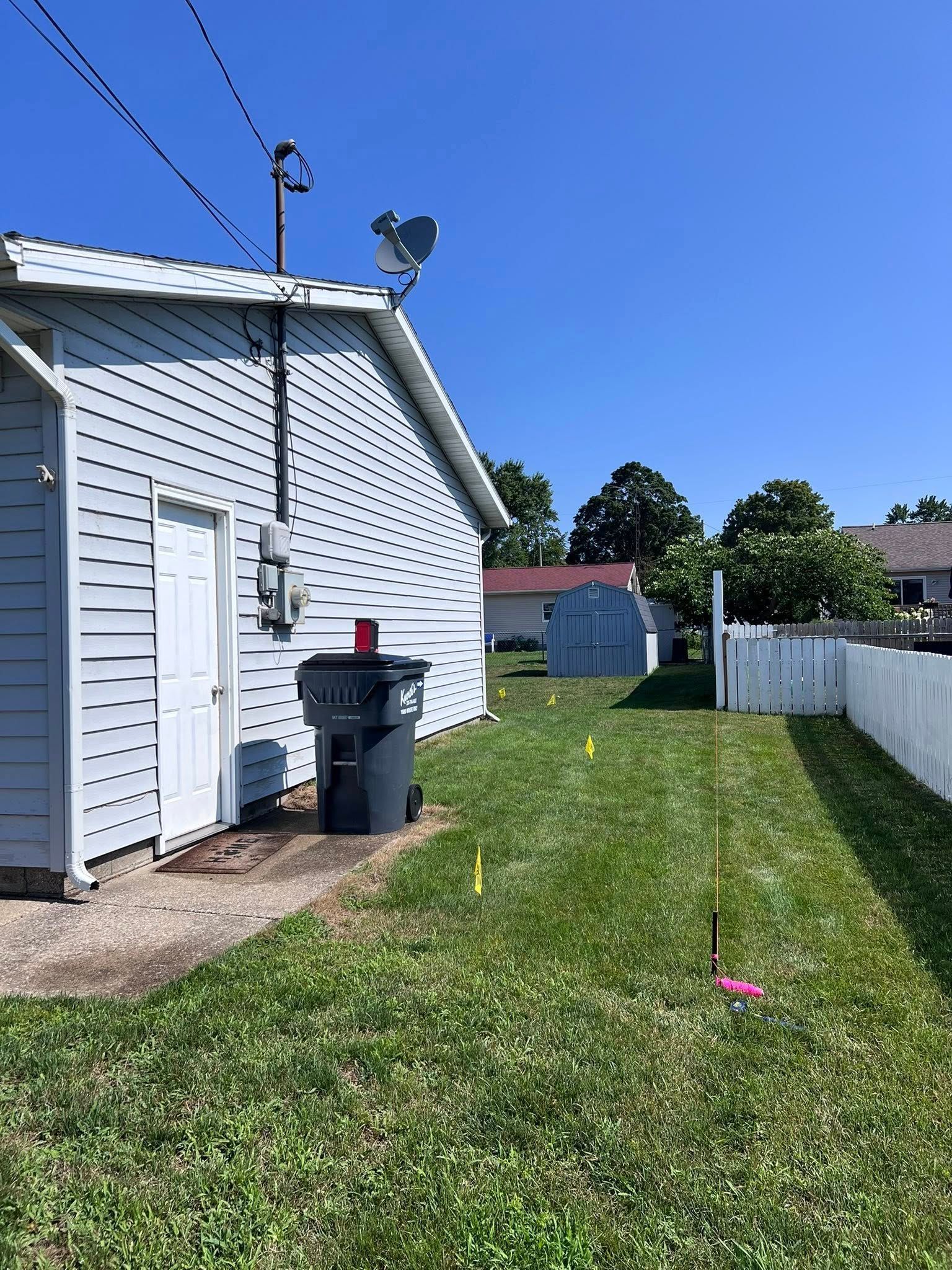 White house side yard with a utility bin, grassy lawn, and white fence under a blue sky.