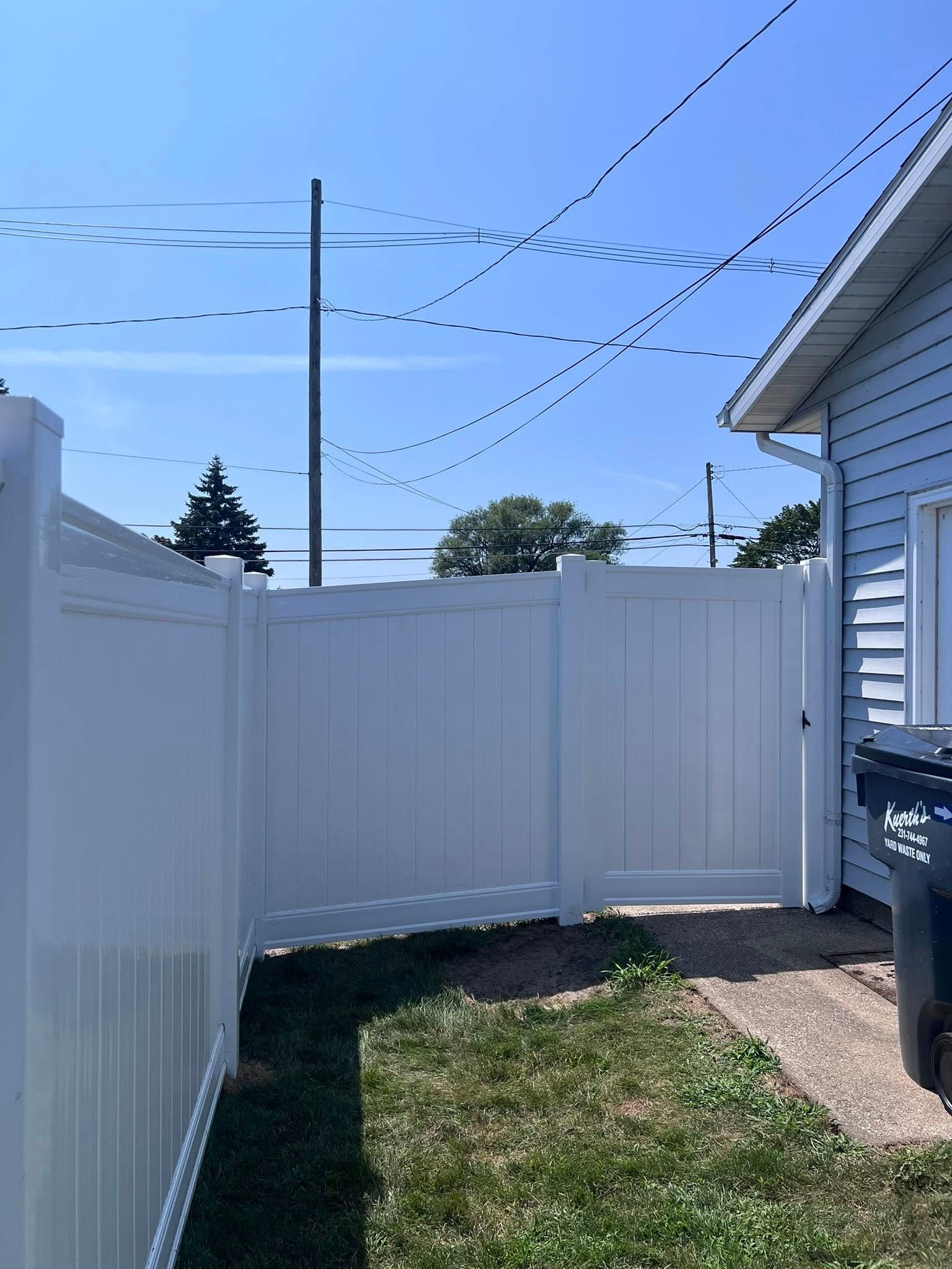 Backyard with white vinyl fence, small grass patch, house siding, and blue sky overhead
