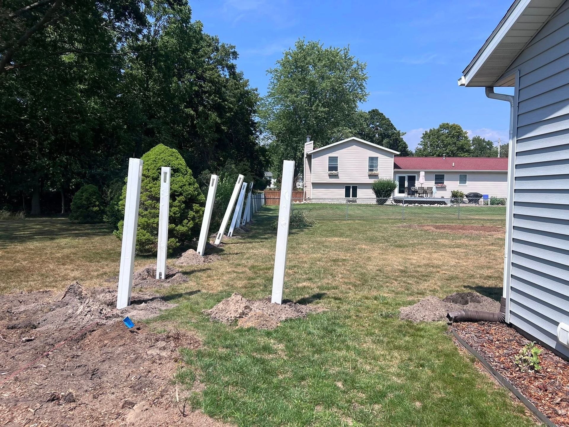 White posts in a grassy yard near a house, with trees and a clear blue sky in the background.