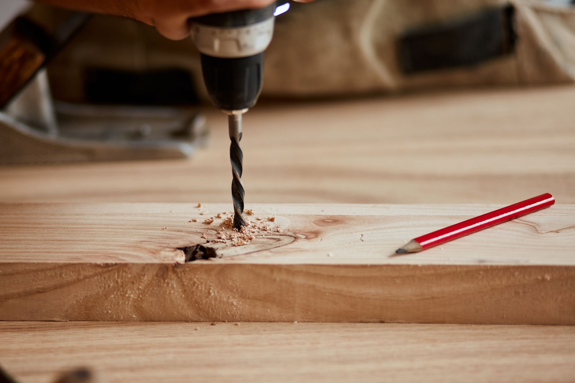 Power drill boring into a wooden board beside a red pencil on a workbench