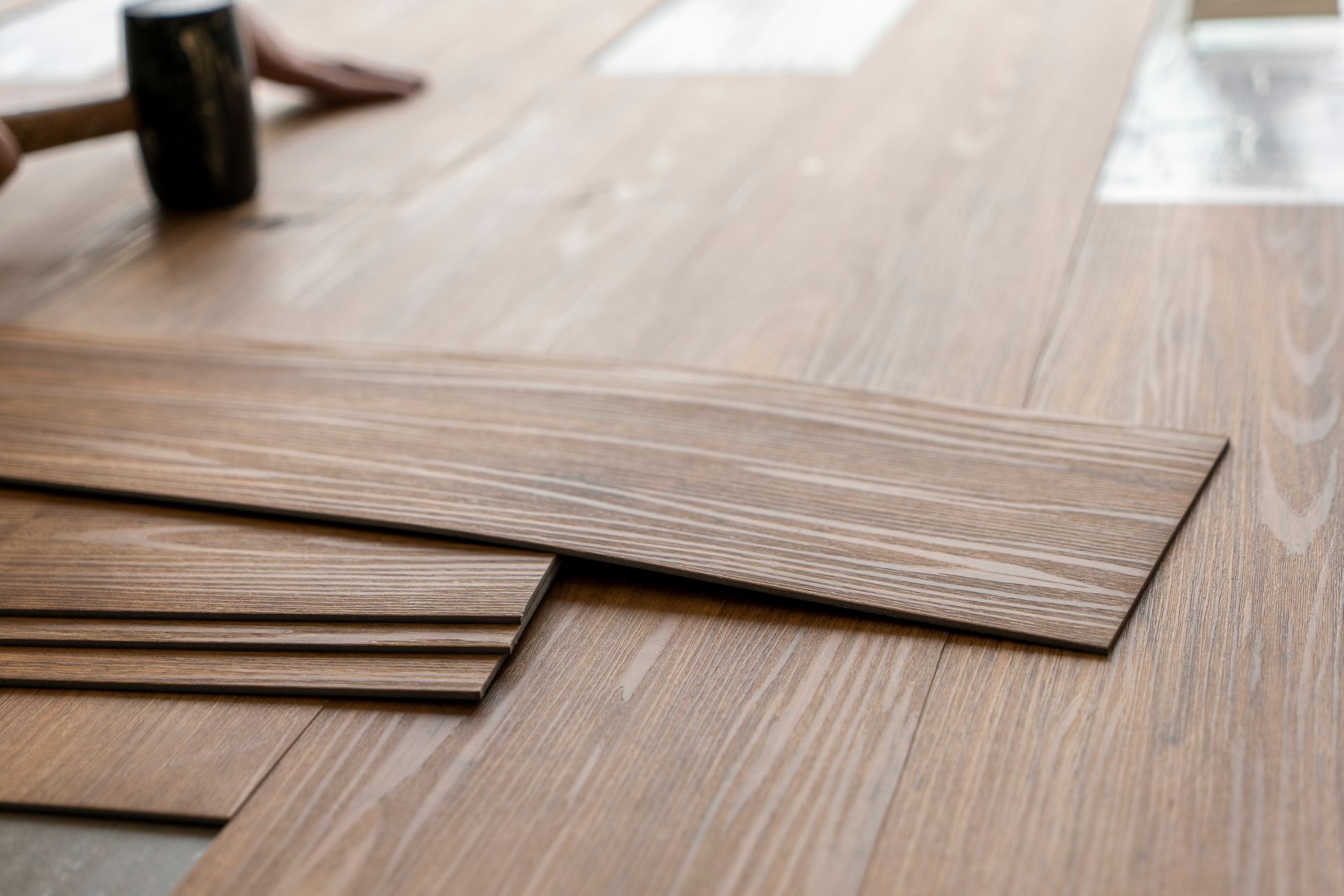 Stacked brown wood-look flooring planks on a light wood floor, with a small can in the background